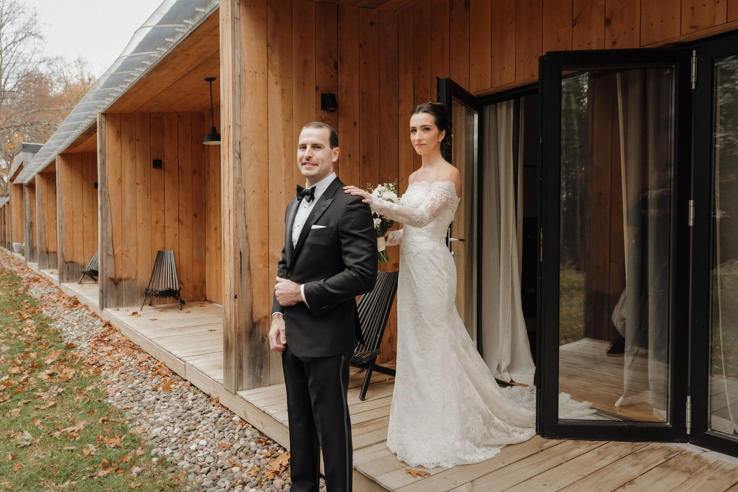 A bride in a white lace wedding dress holding a bouquet stands behind a groom in a black tuxedo, outside a wooden building with glass doors, autumn scenery.