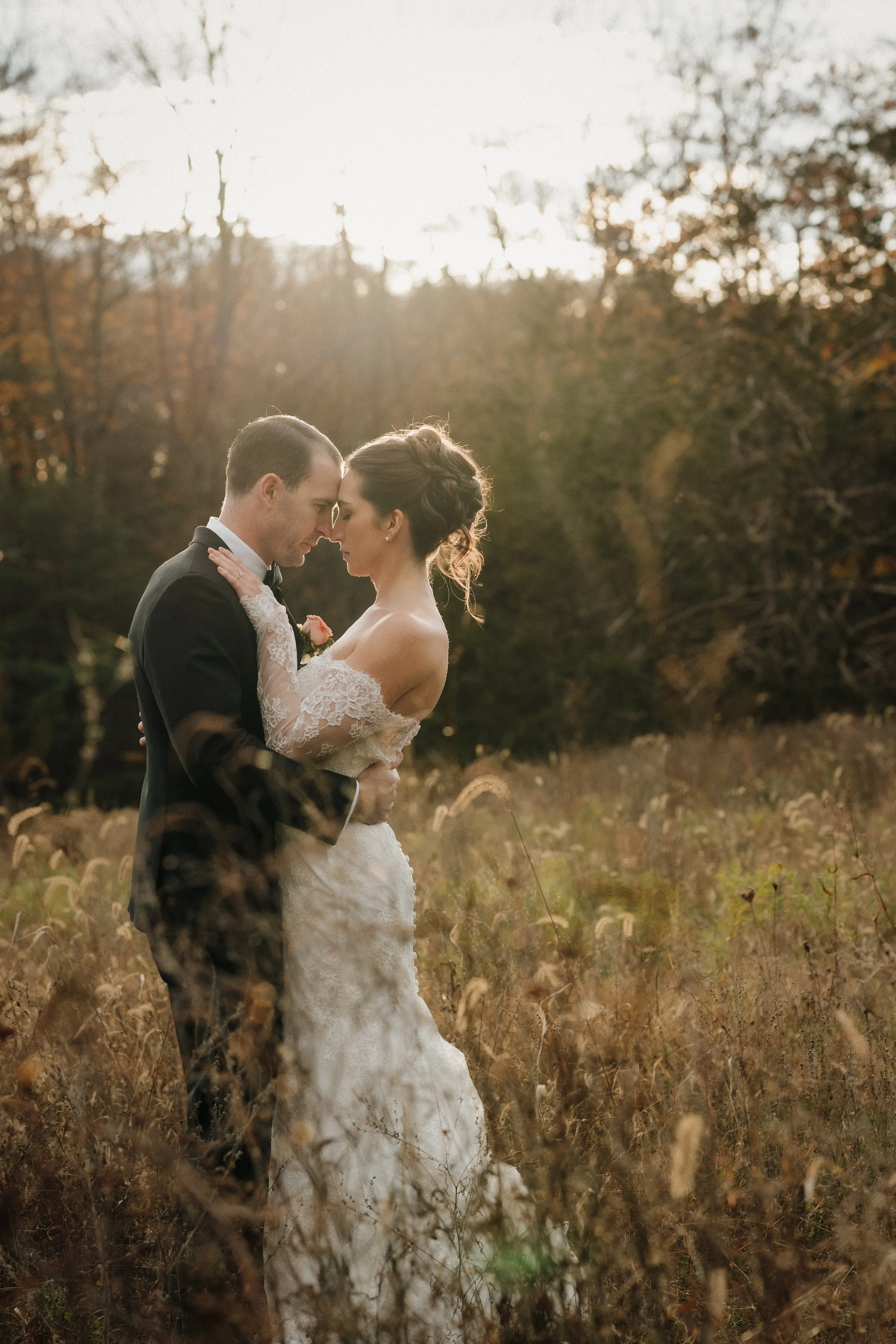 A bride and groom in wedding attire standing close, touching foreheads, in a field with tall grasses and trees in the background during sunset.