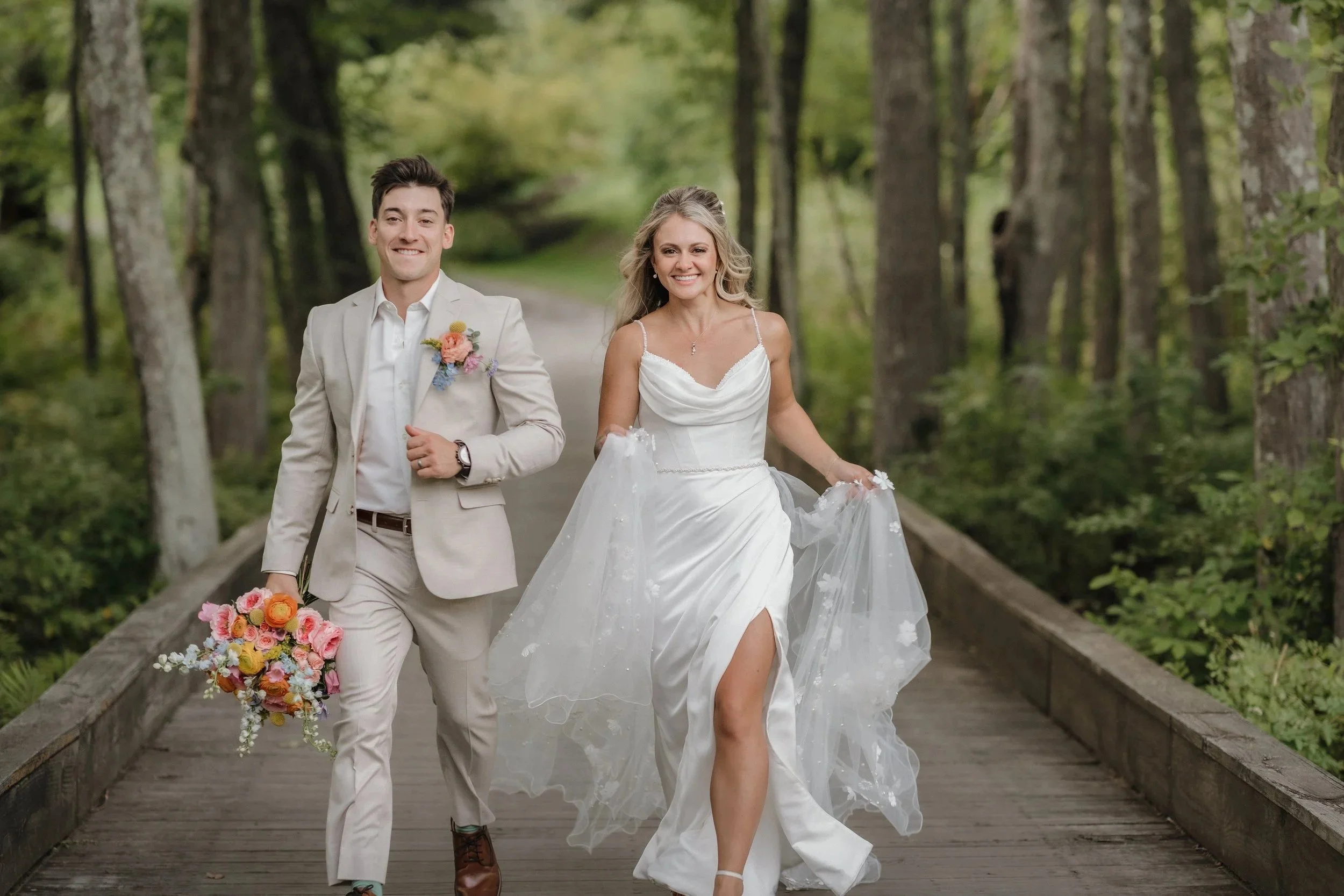 A bride and groom walking on a wooden bridge in a forest, smiling, with the bride holding her dress and the groom holding a colorful bouquet.