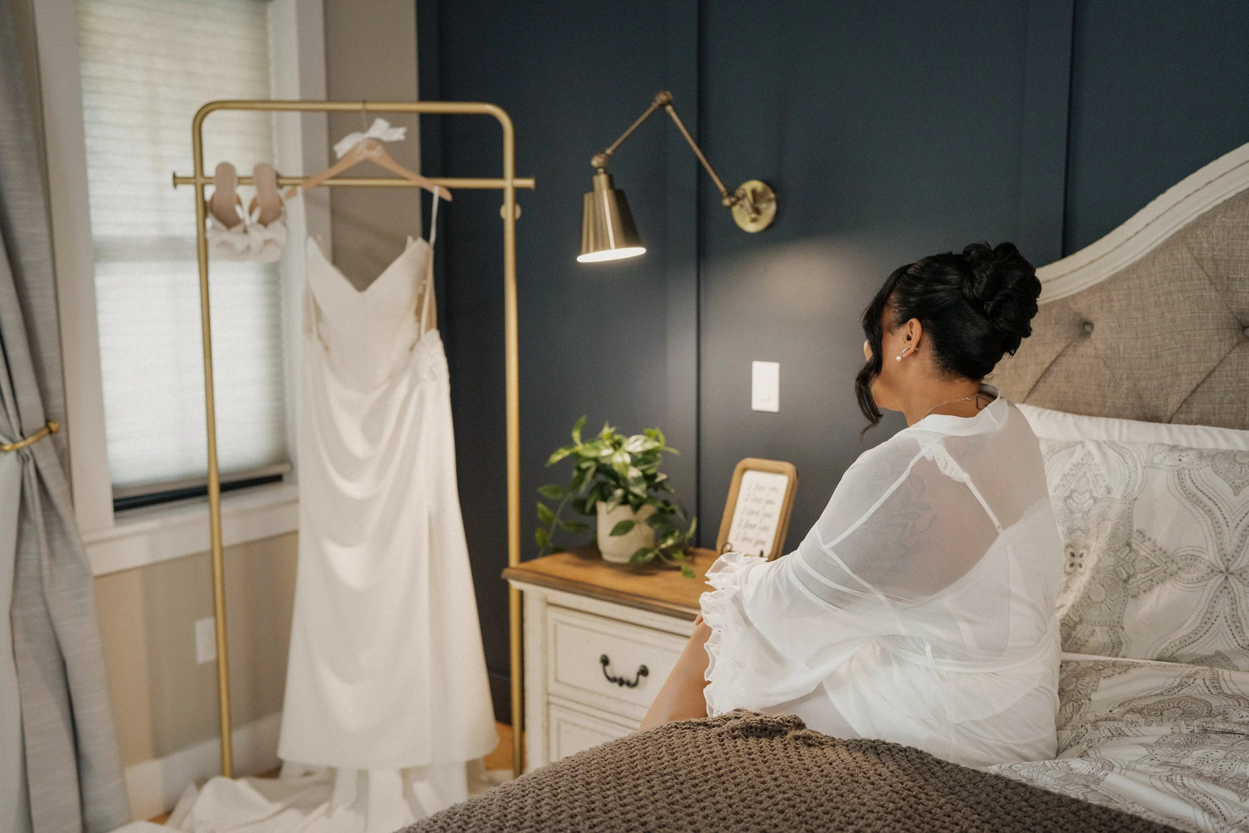 A woman in a white robe sitting on a bed looking at her wedding dress hanging on a gold clothing rack in a bedroom.