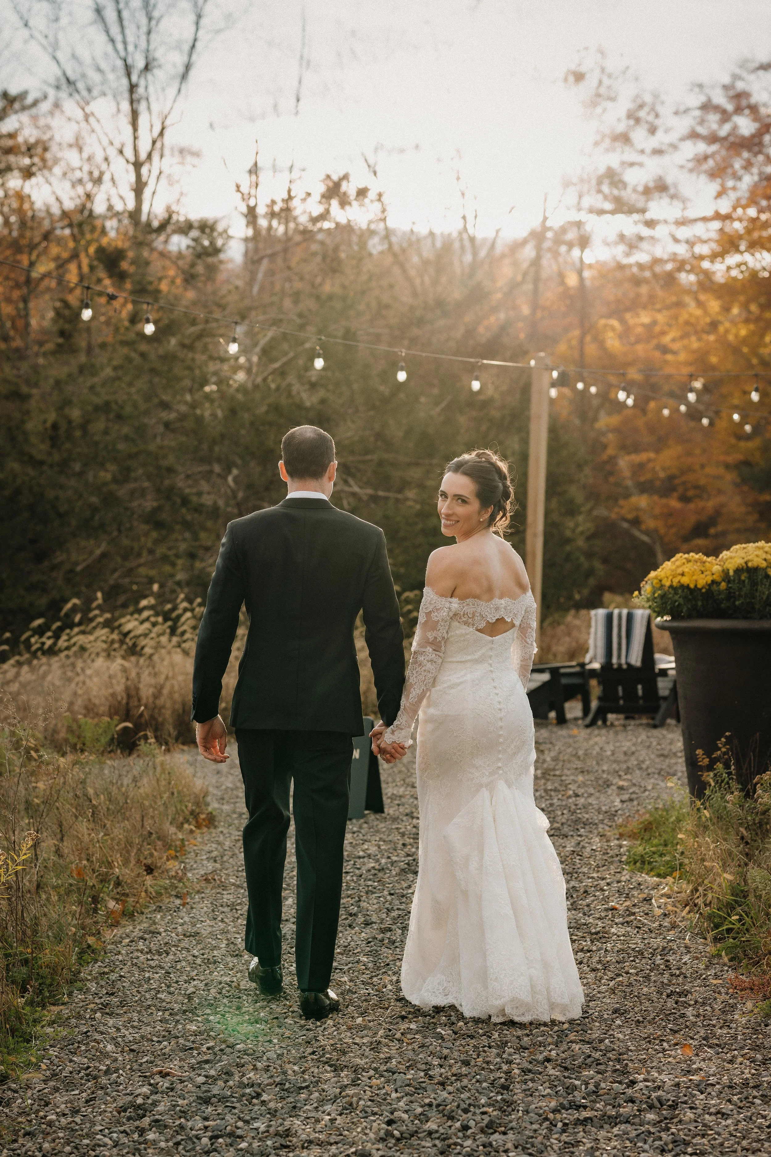 A bride and groom holding hands and walking outdoors on a gravel path during sunset, with trees and string lights overhead.