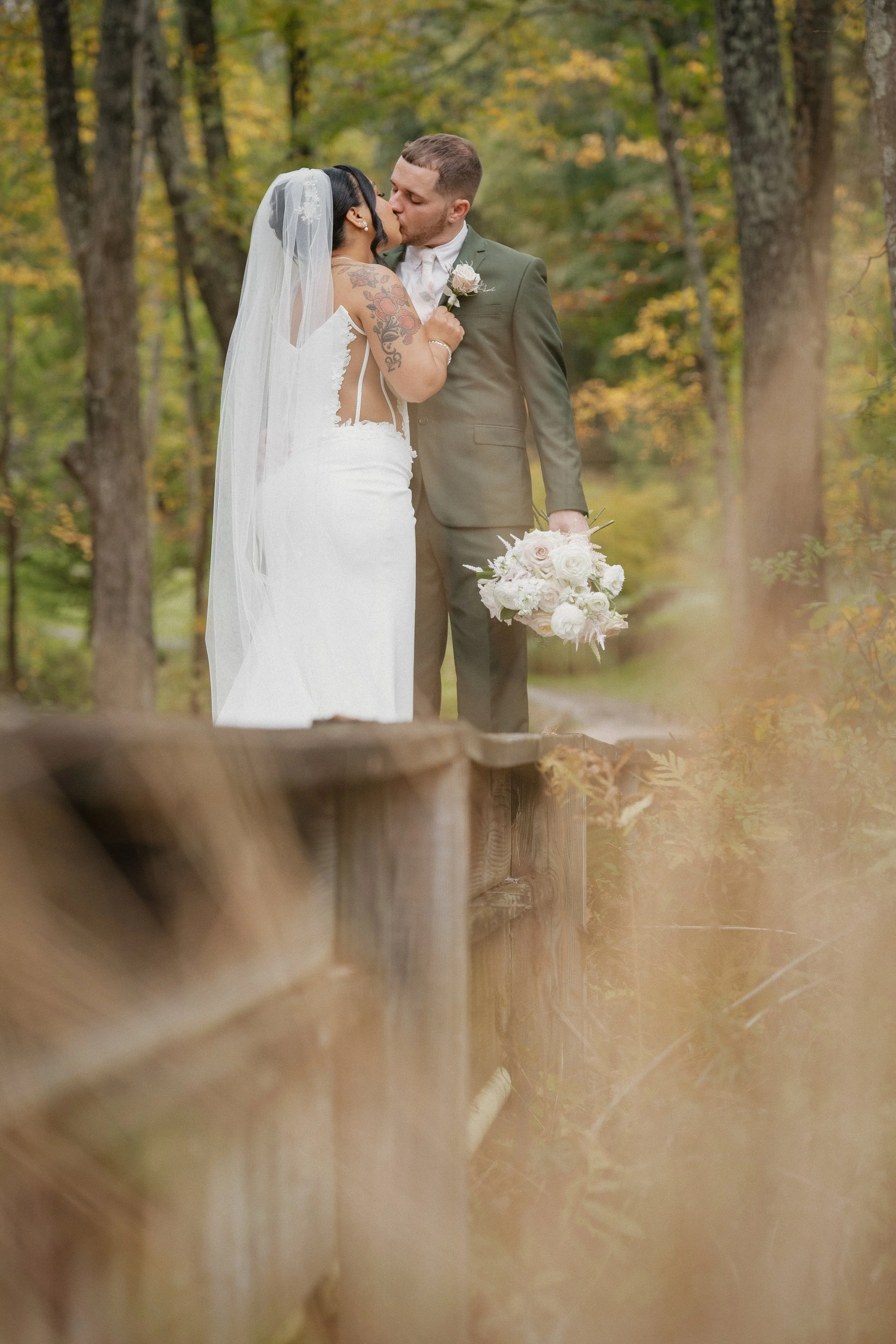 A newly married couple shares a kiss outdoors in a wooded area during fall, with the bride holding a bouquet of white flowers.