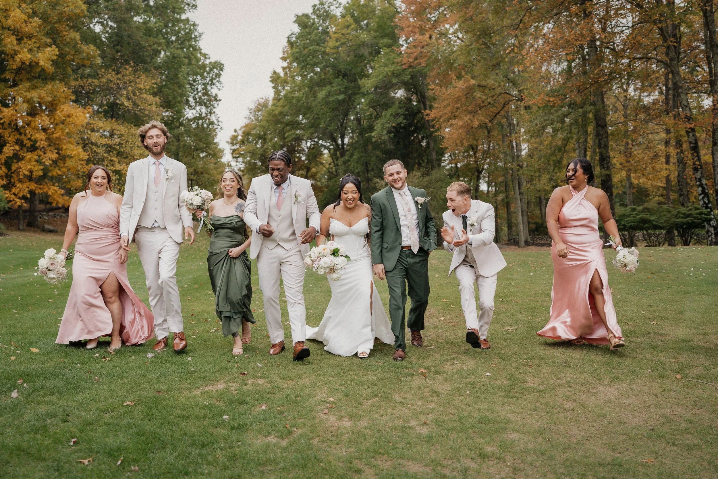 A diverse group of wedding guests, including the bride and groom, walking happily on a grassy field surrounded by autumn trees.
