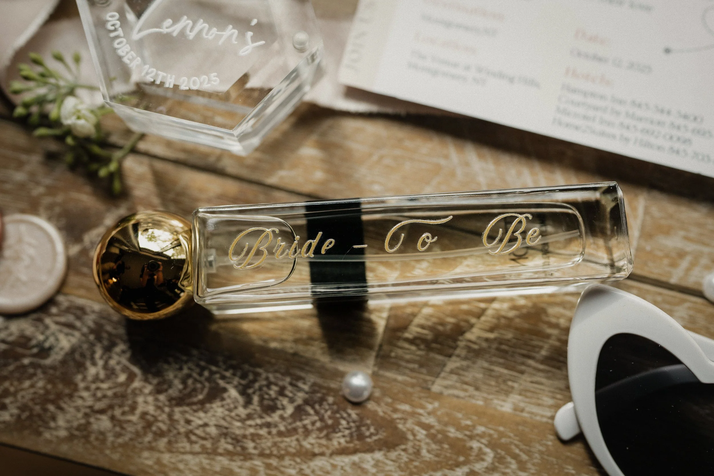 A glass bottle with a gold cap and gold lettering that reads 'Bride - To Be', placed on a wooden surface with miscellaneous wedding-related items nearby, including a clear acrylic block engraved with the name 'Lennon's' and a date, a white flower, a 