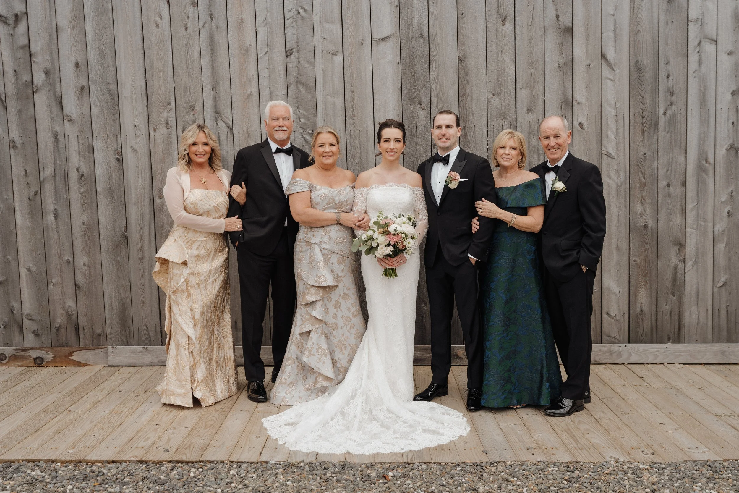 Group of people at a wedding, including a bride in a white lace gown holding a bouquet of flowers, and several guests dressed in formal attire, standing in front of a wooden wall.