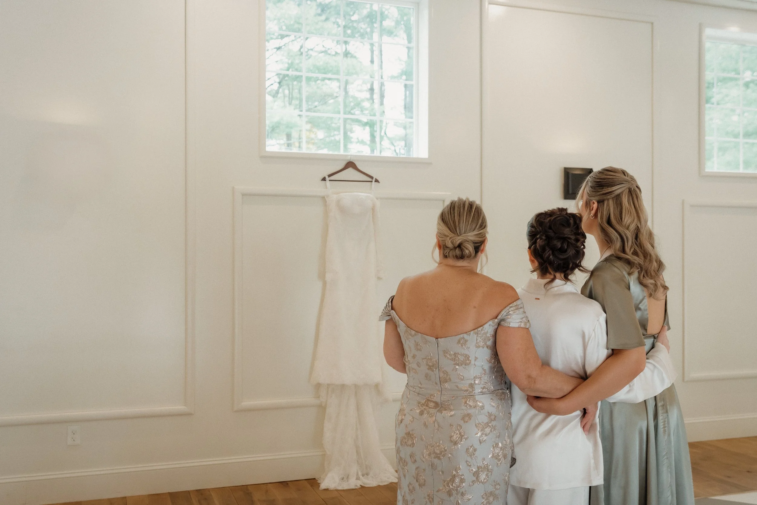 Three women in formal attire standing together in front of a white wall with two windows and a wedding dress hanging on the wall, likely at a wedding preparation.