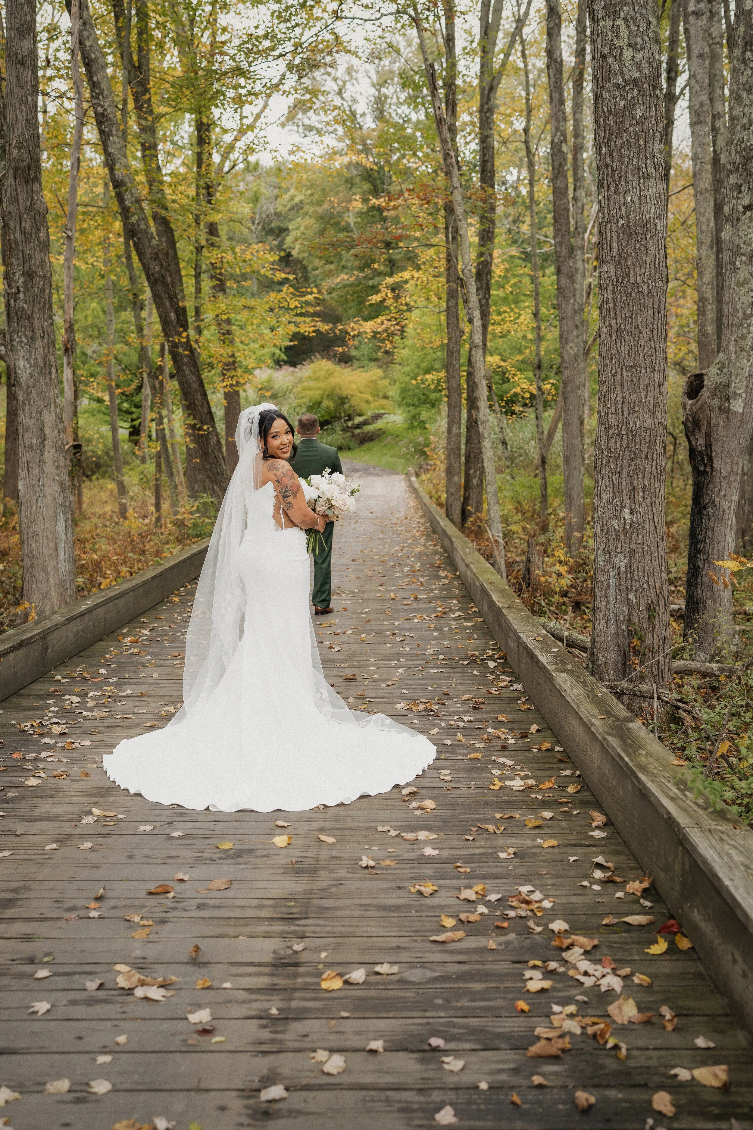 A bride in a white wedding dress holding a bouquet, standing on a wooden bridge in a forest during autumn, with a groom walking ahead.