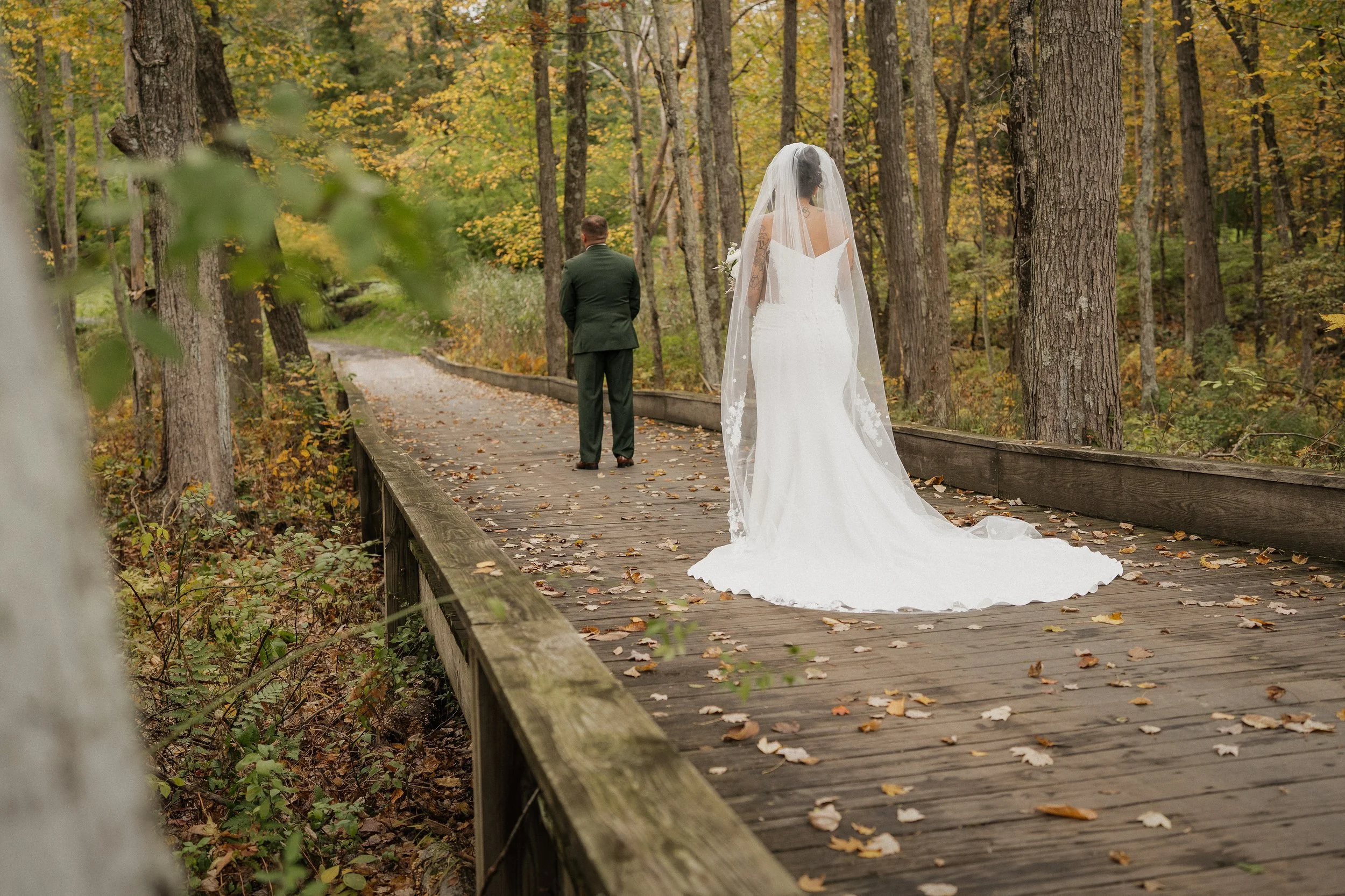 A bride in a white wedding gown and veil standing on a wooden bridge in an autumn forest, with a man in a dark suit walking ahead on the same bridge.