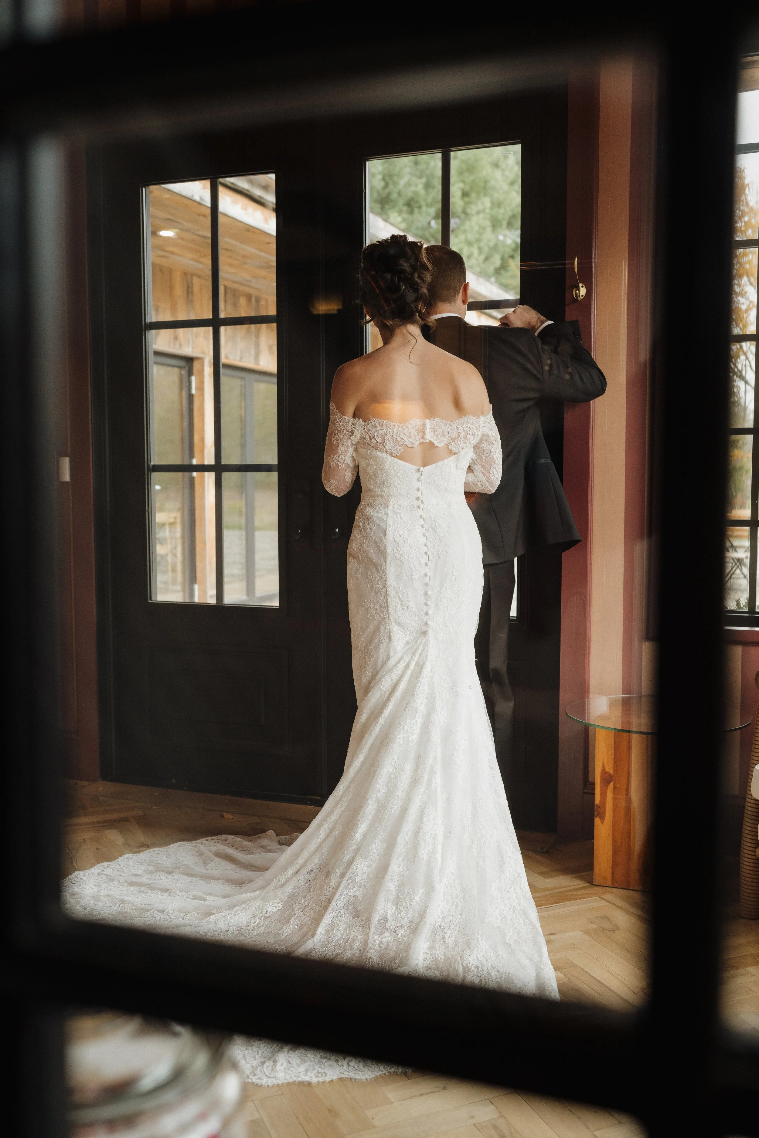Bride in an off-shoulder lace wedding dress standing by a window, groom in a black suit adjusting his cufflink inside a warmly lit room.