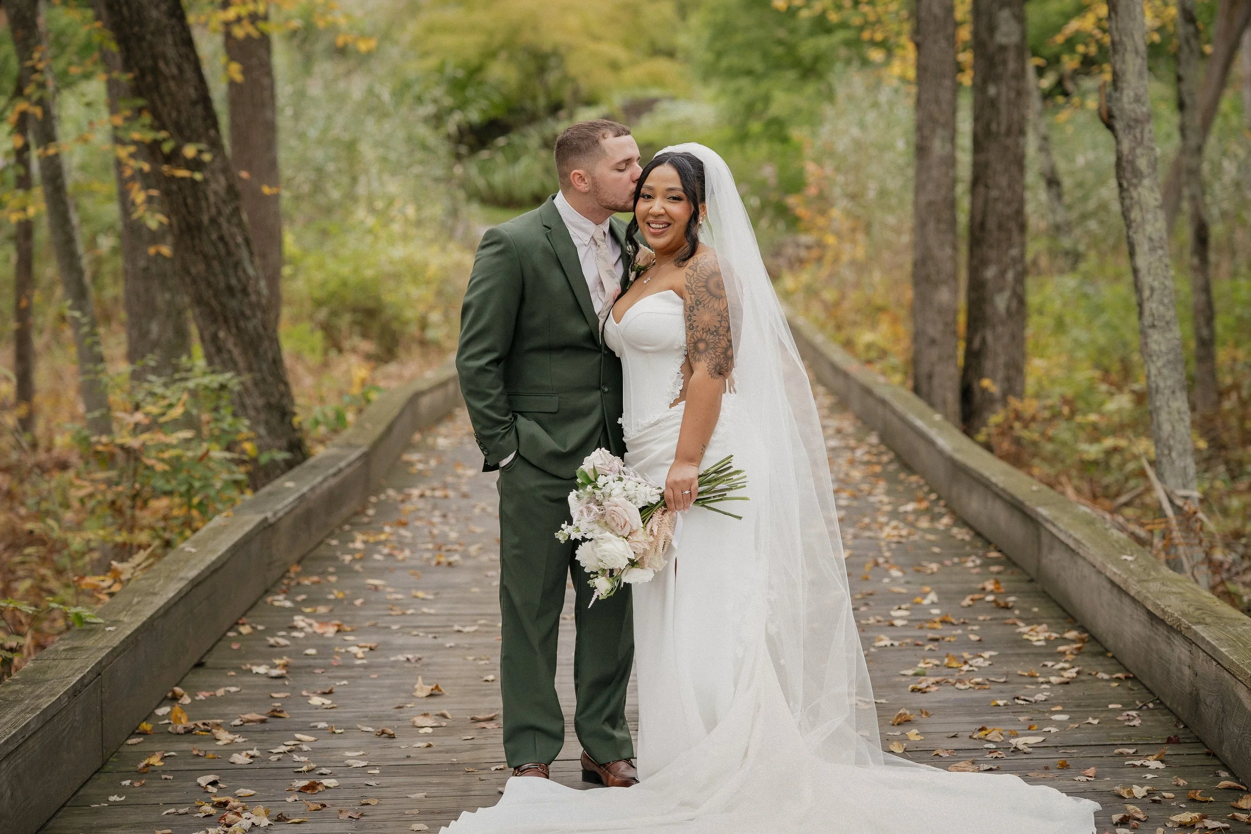 A newlywed couple standing on a wooden bridge in a forest, with the groom kissing the bride's temple as she smiles, holding a bouquet of flowers, during autumn.