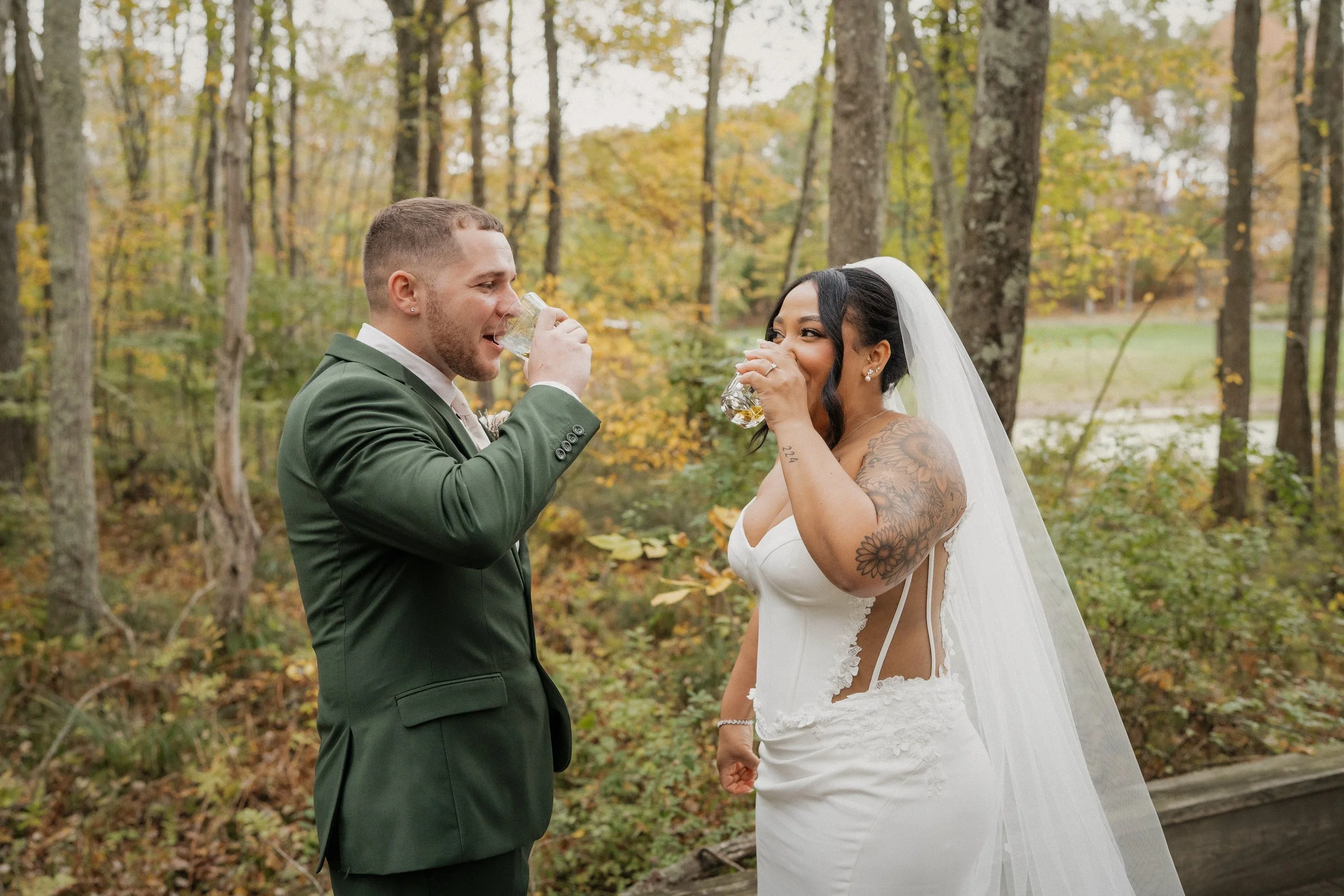 Bride and groom toast with drinks outdoors during fall, surrounded by trees with autumn leaves.