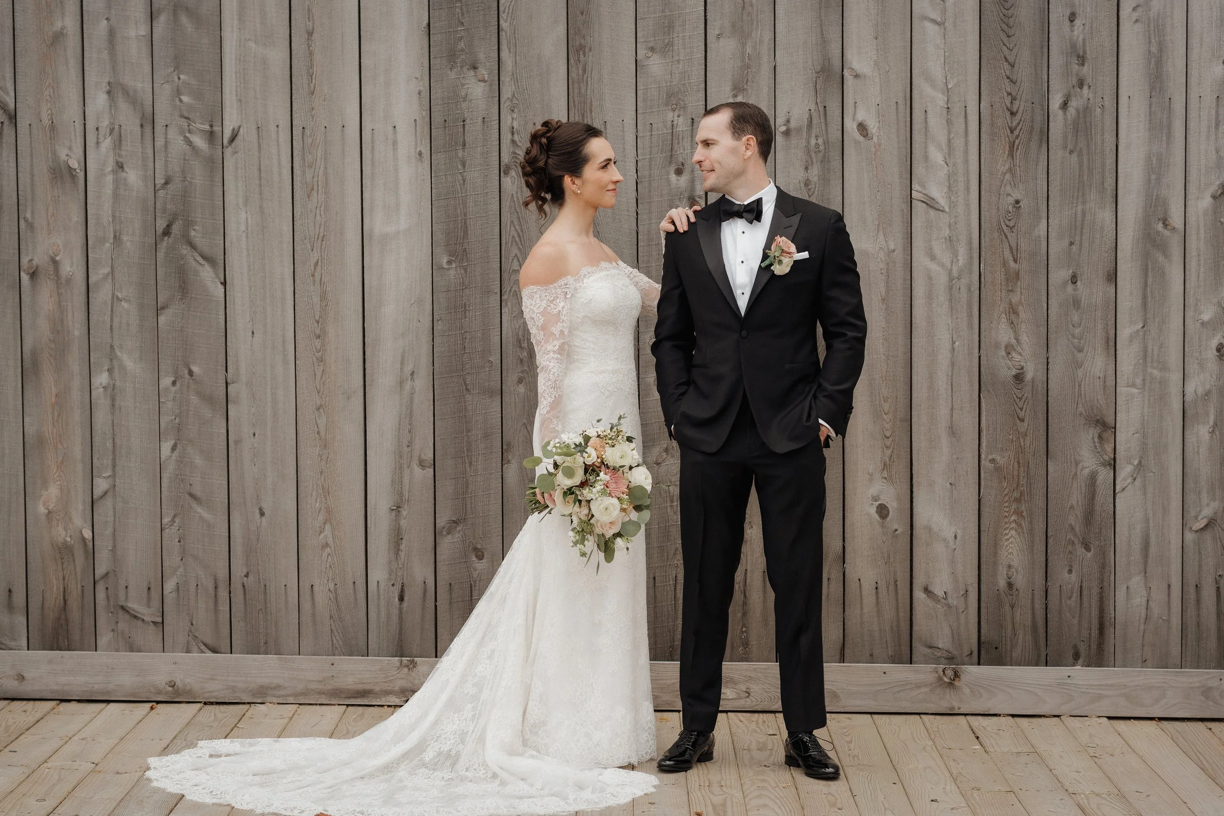 A bride and groom stand facing each other against a wooden background. The bride is holding a bouquet of flowers and wears a white lace wedding gown with off-shoulder sleeves. The groom is dressed in a black tuxedo with a white shirt, black bow tie, 