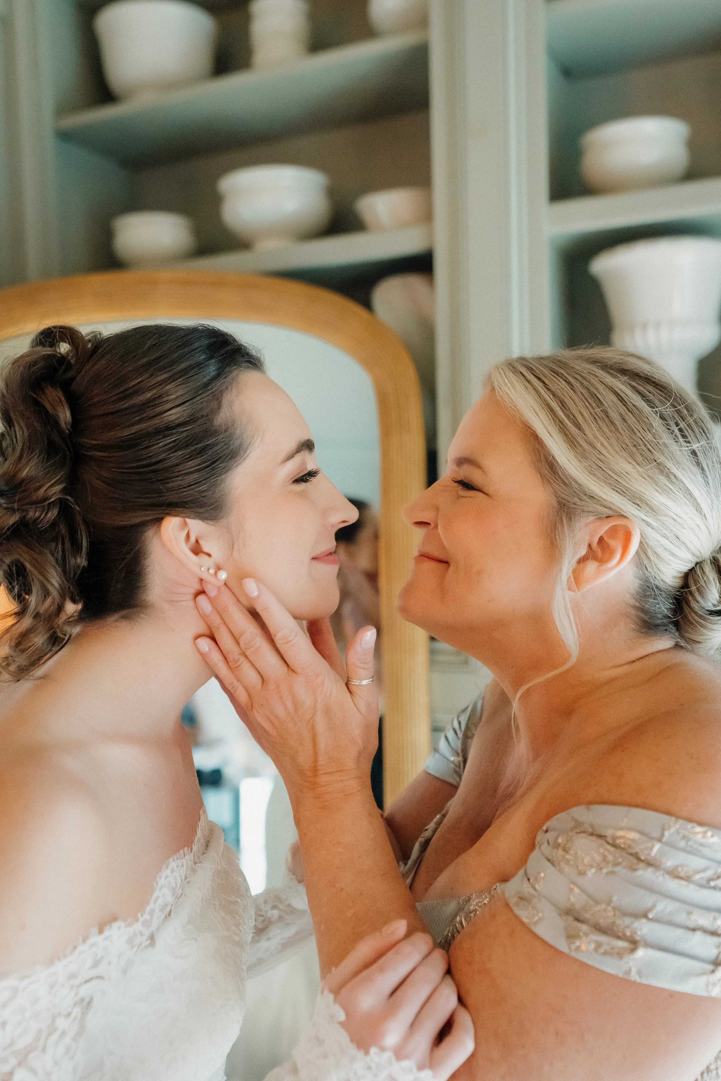 A woman, likely the bride, smiling as she looks at an older woman, possibly her mother, who holds her face gently. The scene is warm and intimate, with a mirror and shelves with white pottery in the background.