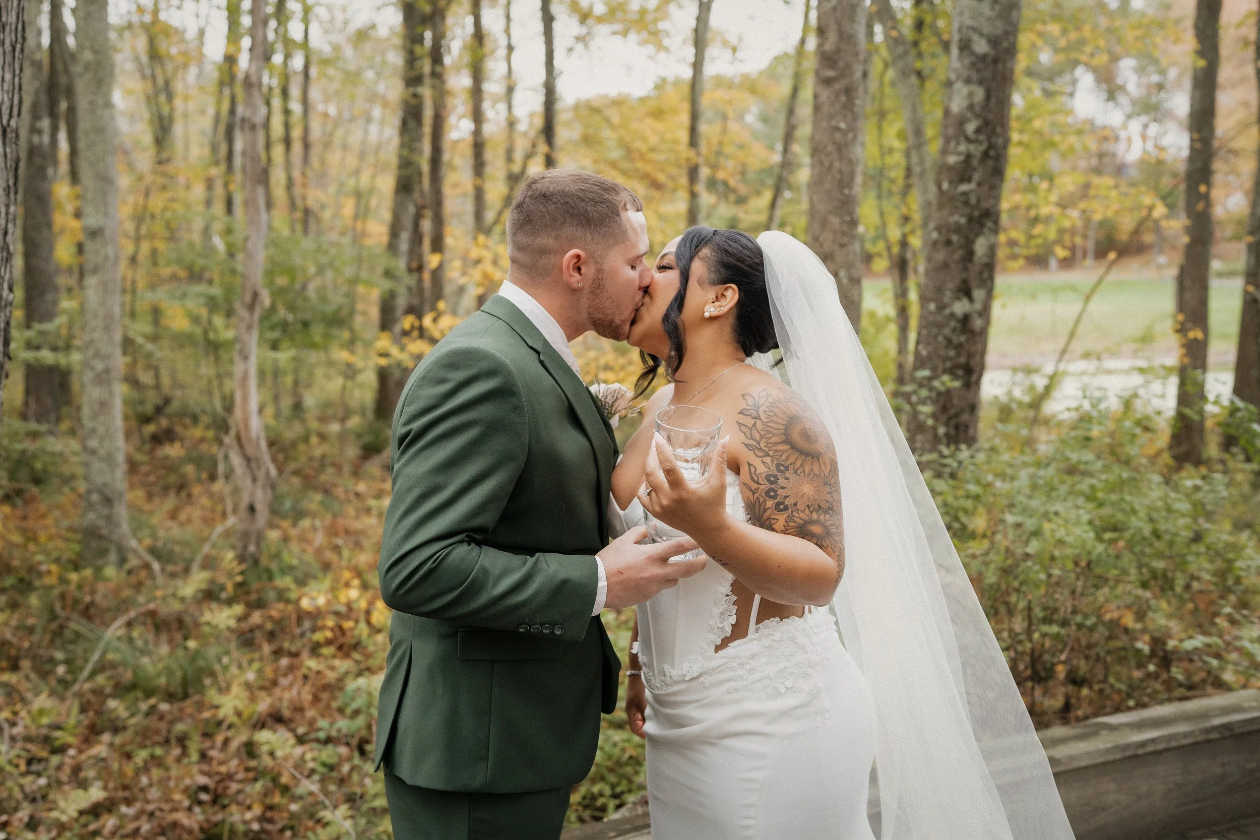 A newlywed couple sharing a kiss in a wooded outdoor setting during autumn. The groom is wearing a green suit and the bride has a veil, floral tattoo on her arm, and is holding a glass of water.