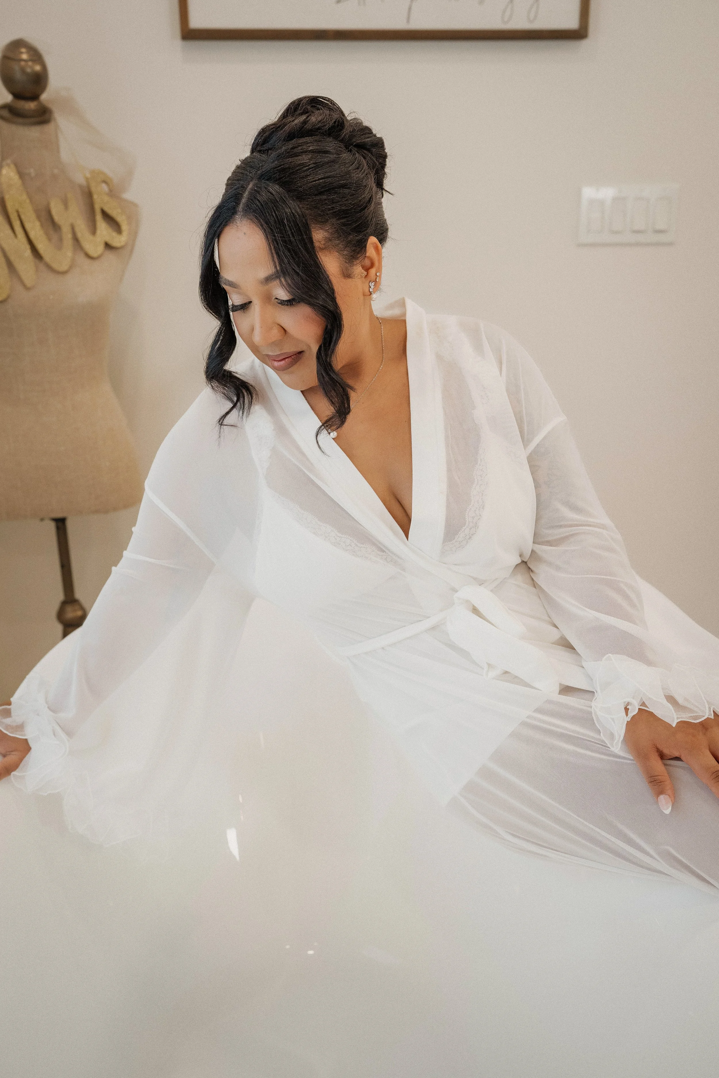 A woman in a white robe sitting on a white bed, looking down with a gentle smile, with a dress form and wall art in the background.