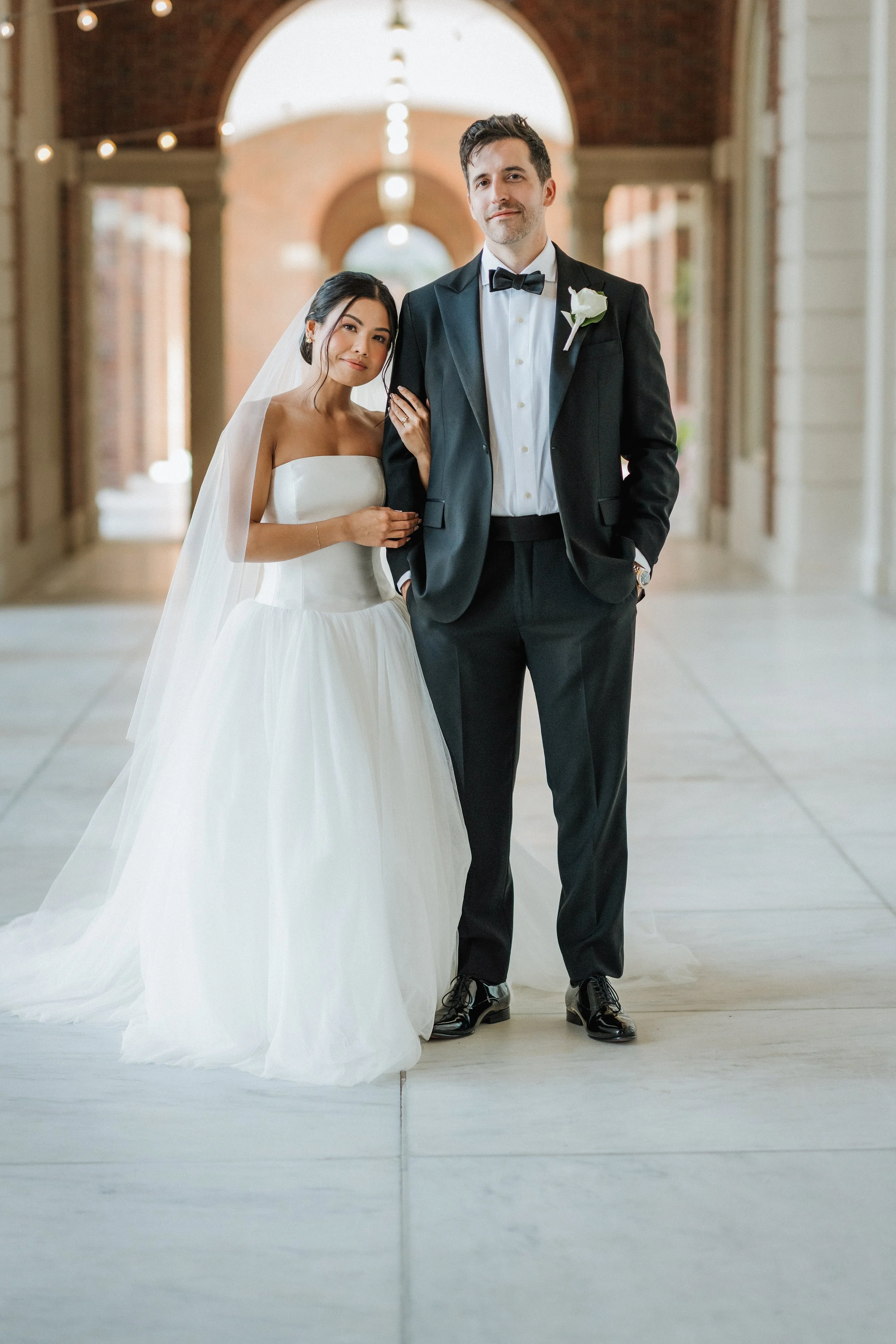 A bride and groom standing inside a covered walkway, with the bride wearing a white wedding gown and veil, and the groom wearing a black tuxedo with a bow tie and boutonniere.