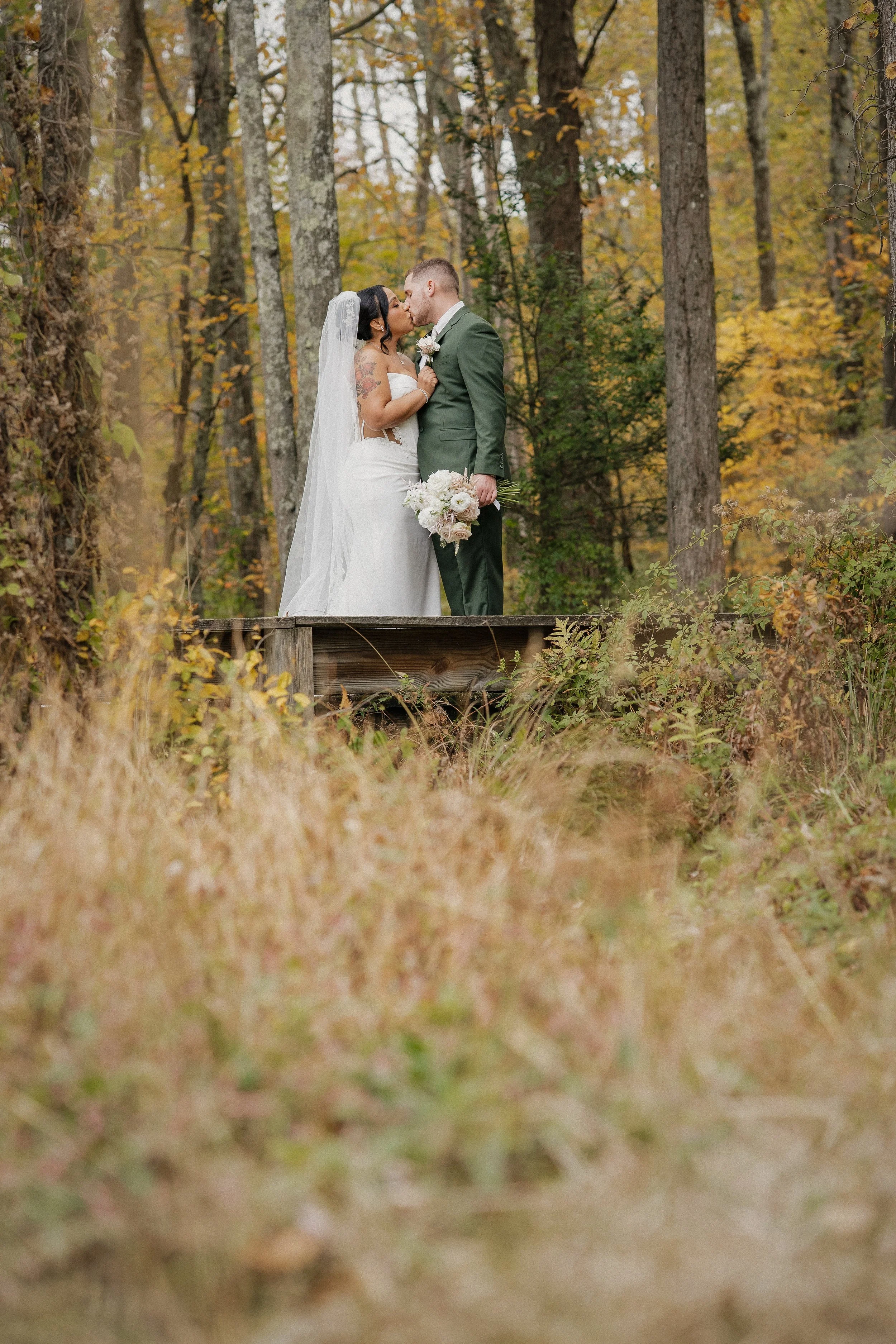 A bride and groom kissing on a wooden platform in a forest with autumn foliage, the bride holding a bouquet of white and pink flowers.