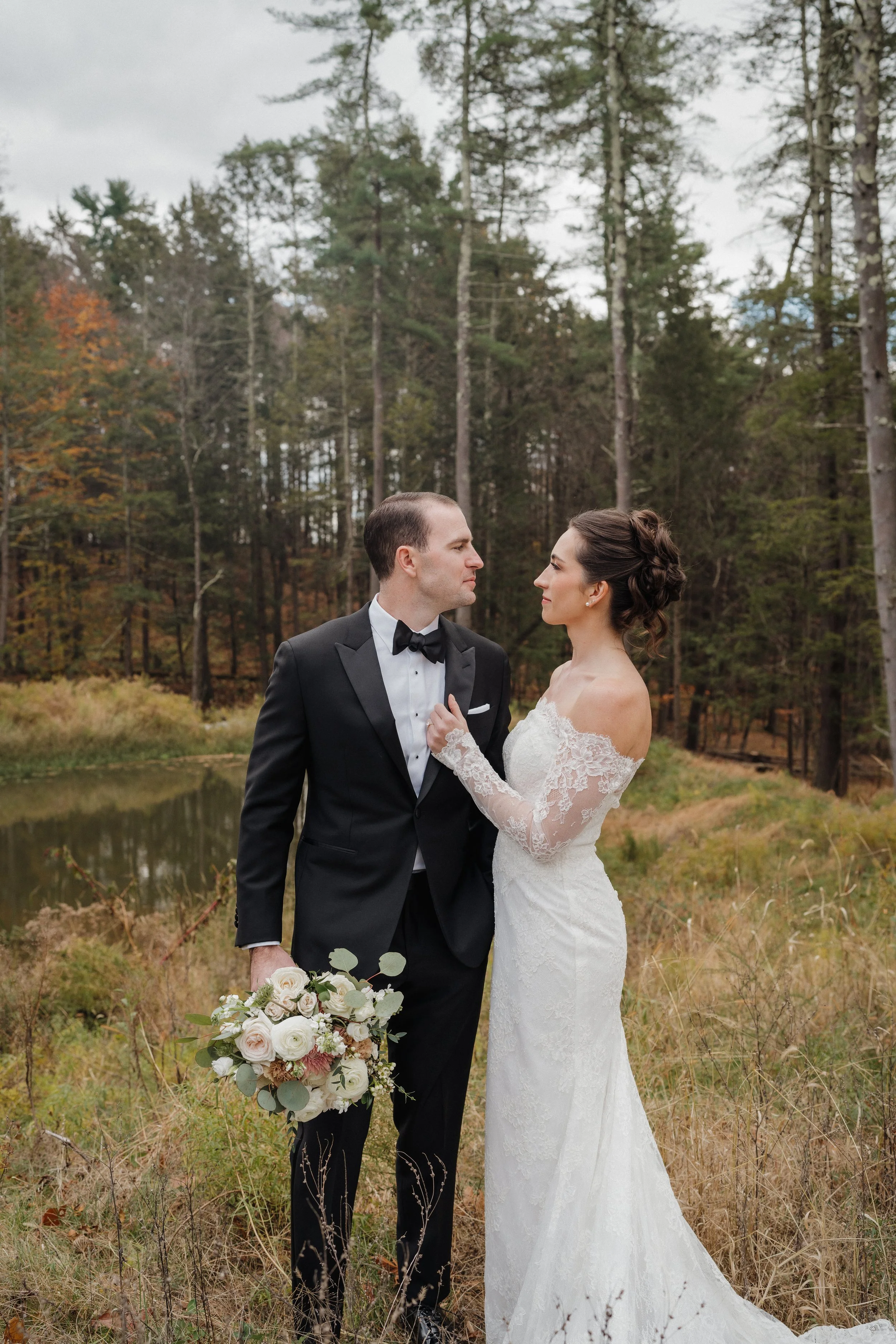 A bride and groom stand facing each other outdoors in a wooded area, with the bride holding a bouquet of flowers.