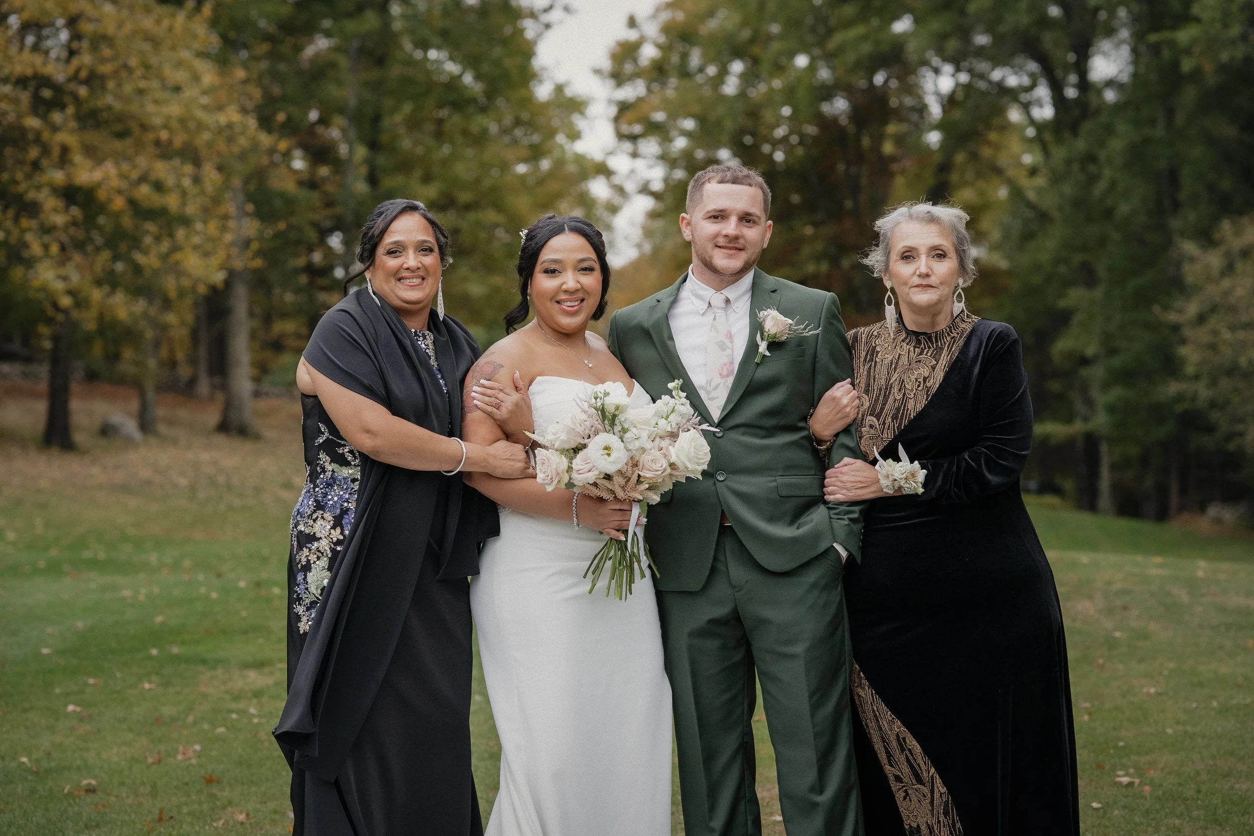 A wedding party outdoors with four people. The bride, holding a bouquet, stands next to a groom in a green suit. Two women, one in a black dress and another in a black velvet dress with gold accents, are beside them. The background features trees wit