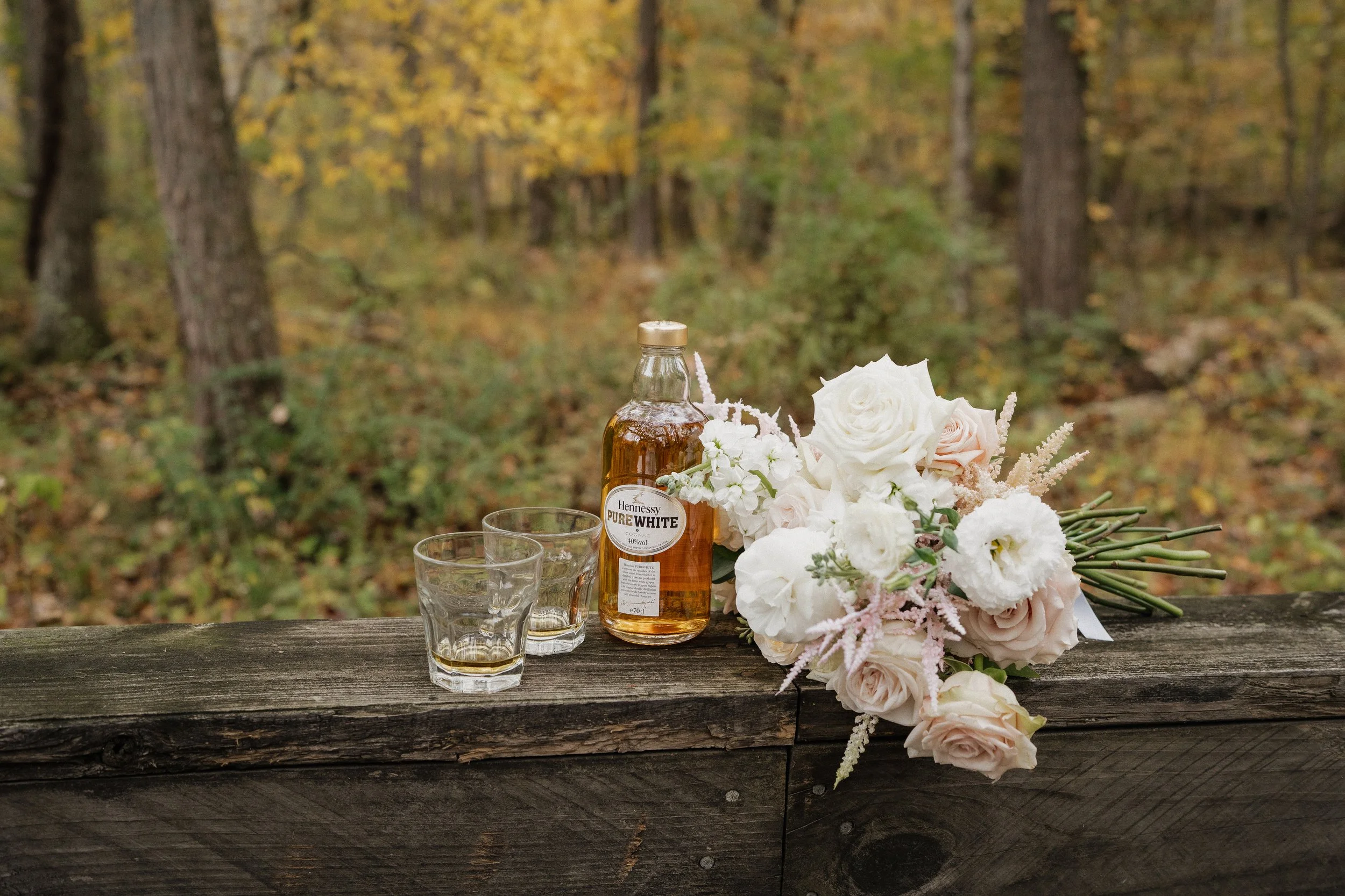 A bottle of Hennessy Pure White whiskey, two empty glasses, and a bouquet of white and pale pink flowers resting on a wooden railing outdoors in a wooded area with fall foliage.