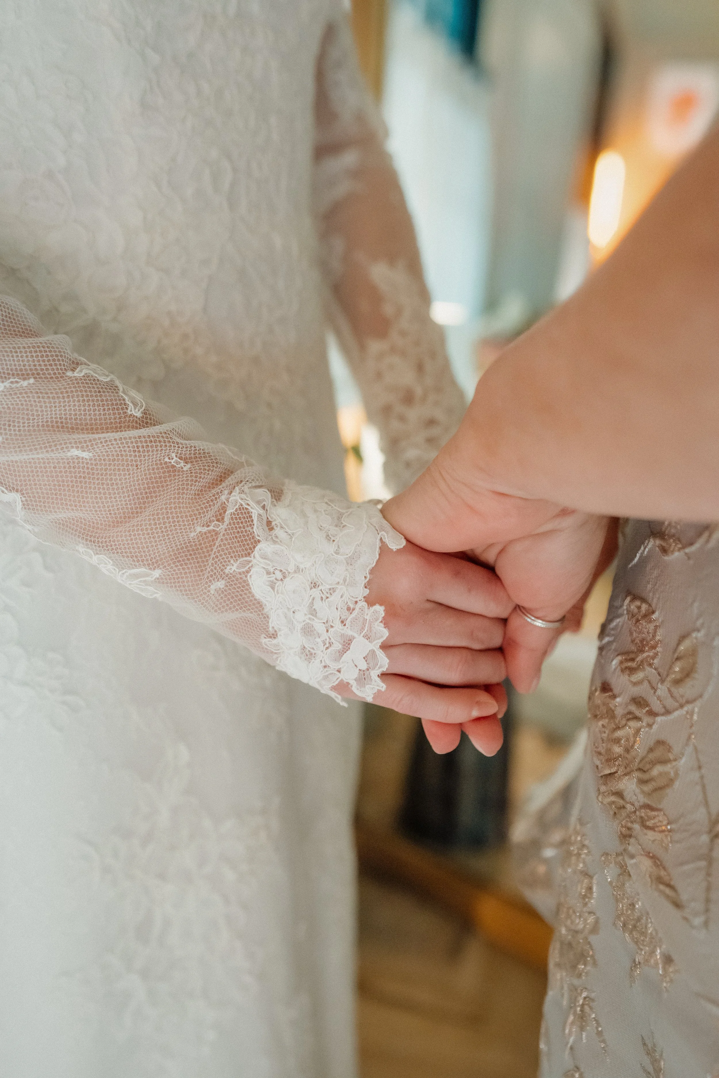 A close-up view of two people holding hands, one wearing a lace sleeve of a wedding dress and the other wearing a ring.