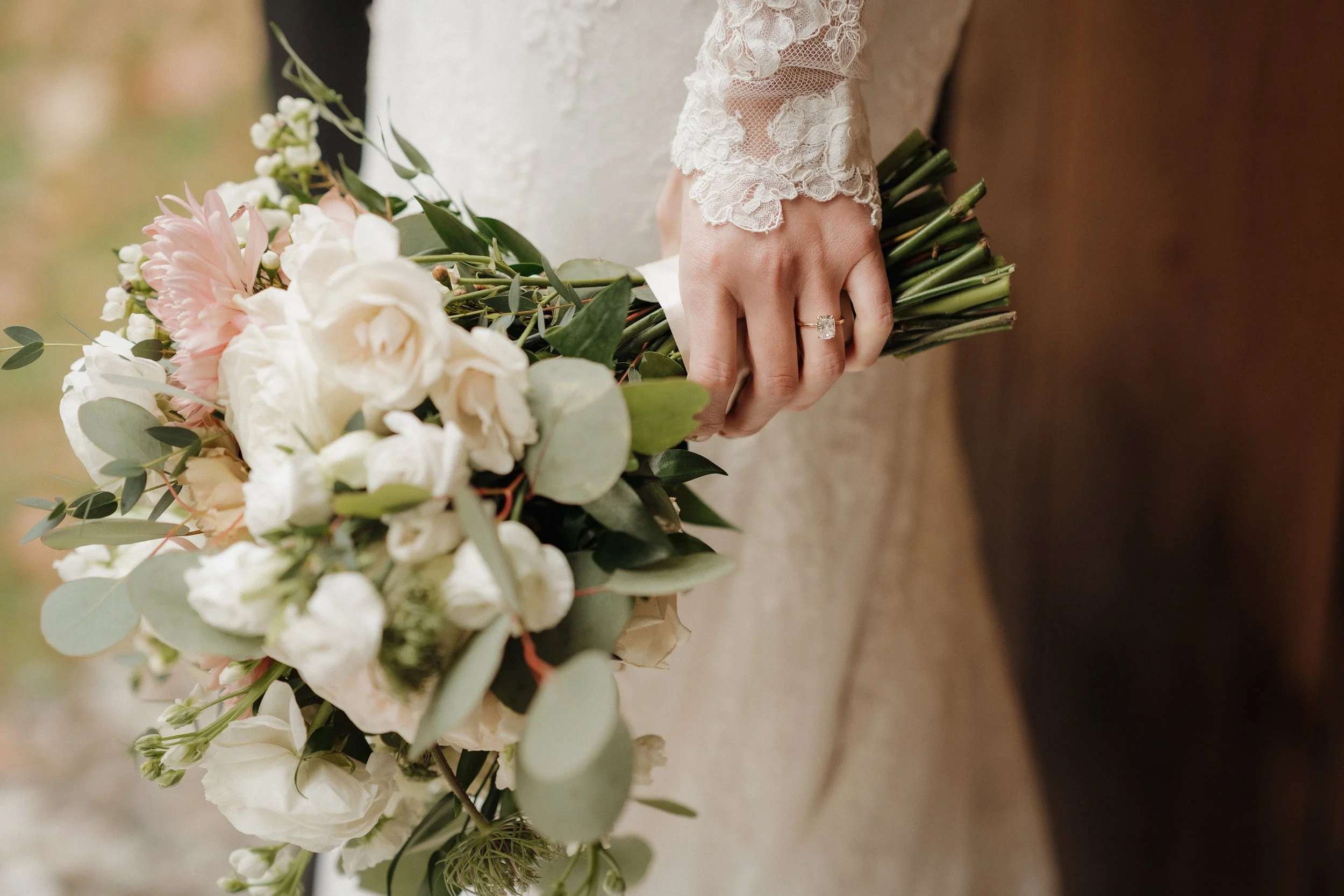 A bride holding a bouquet of white and pink flowers with green leaves, wearing a lace dress with a ring on her finger.