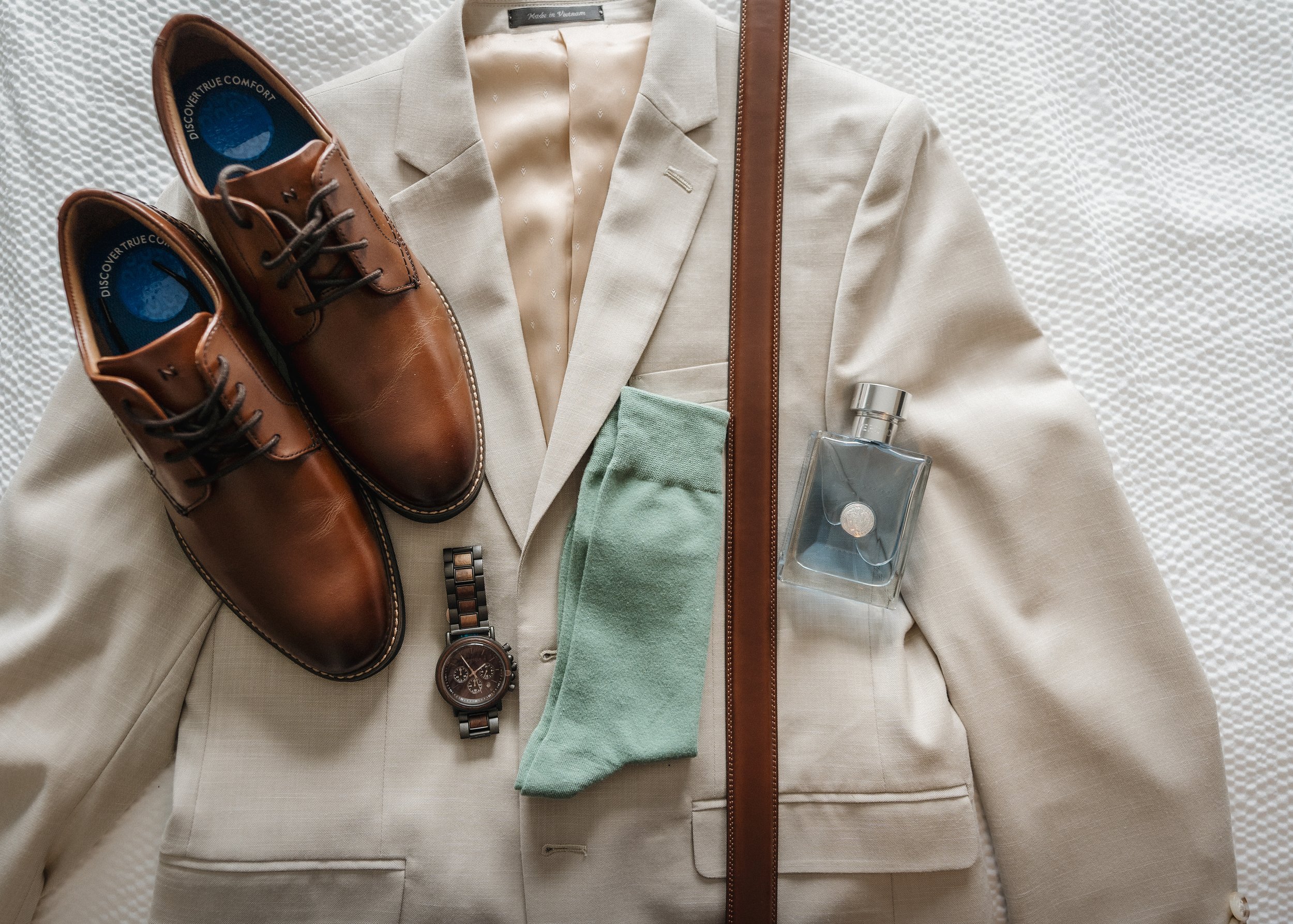 A beige blazer, brown leather dress shoes, a silver wristwatch, a bottle of cologne, a green sock, and a brown leather belt are arranged on a white textured surface, suggesting men's fashion accessories and clothing.