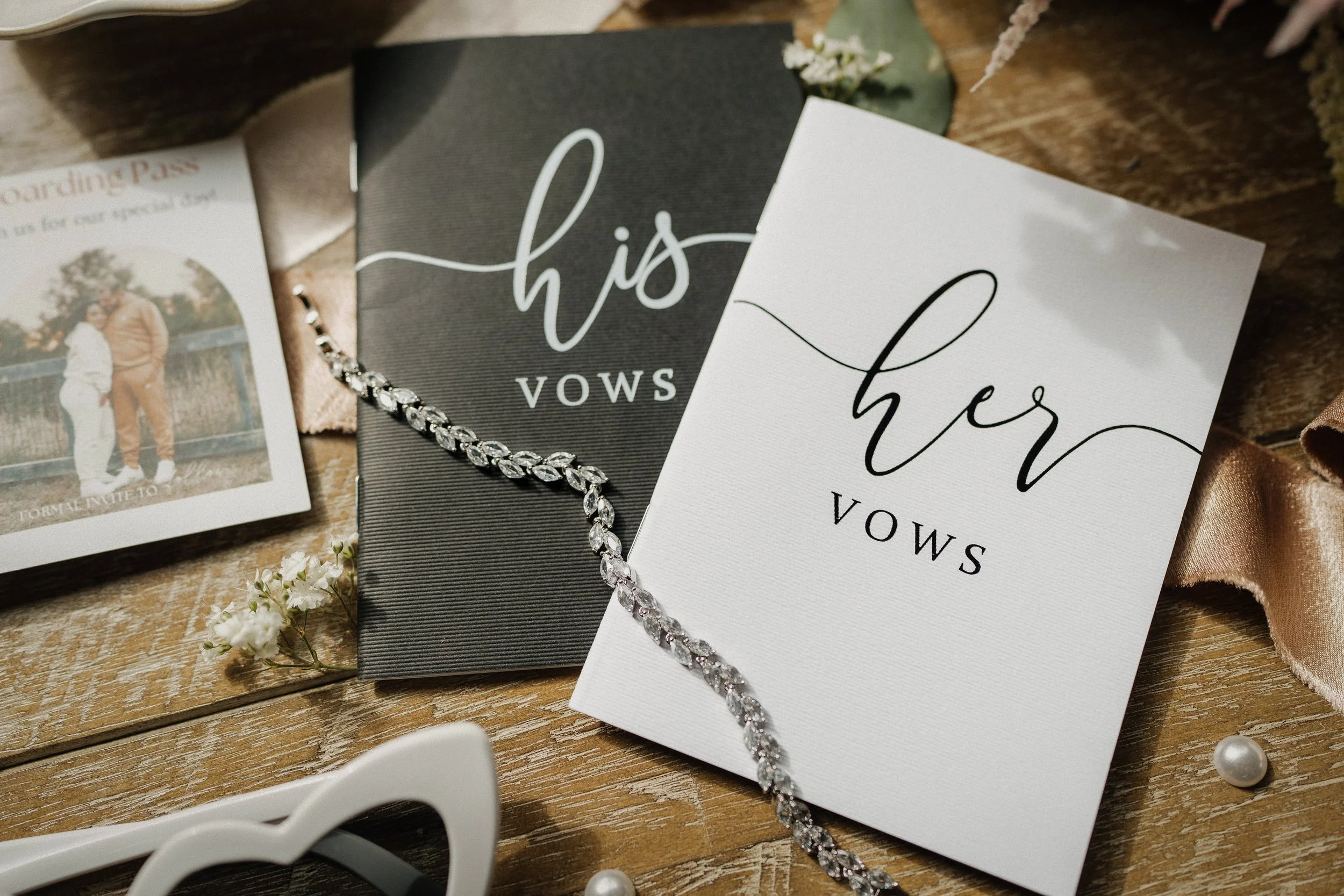 Wedding vow cards with 'his vows' in cursive and 'her vows' in cursive, a diamond necklace, a wedding photo, and pearls on a wooden surface.