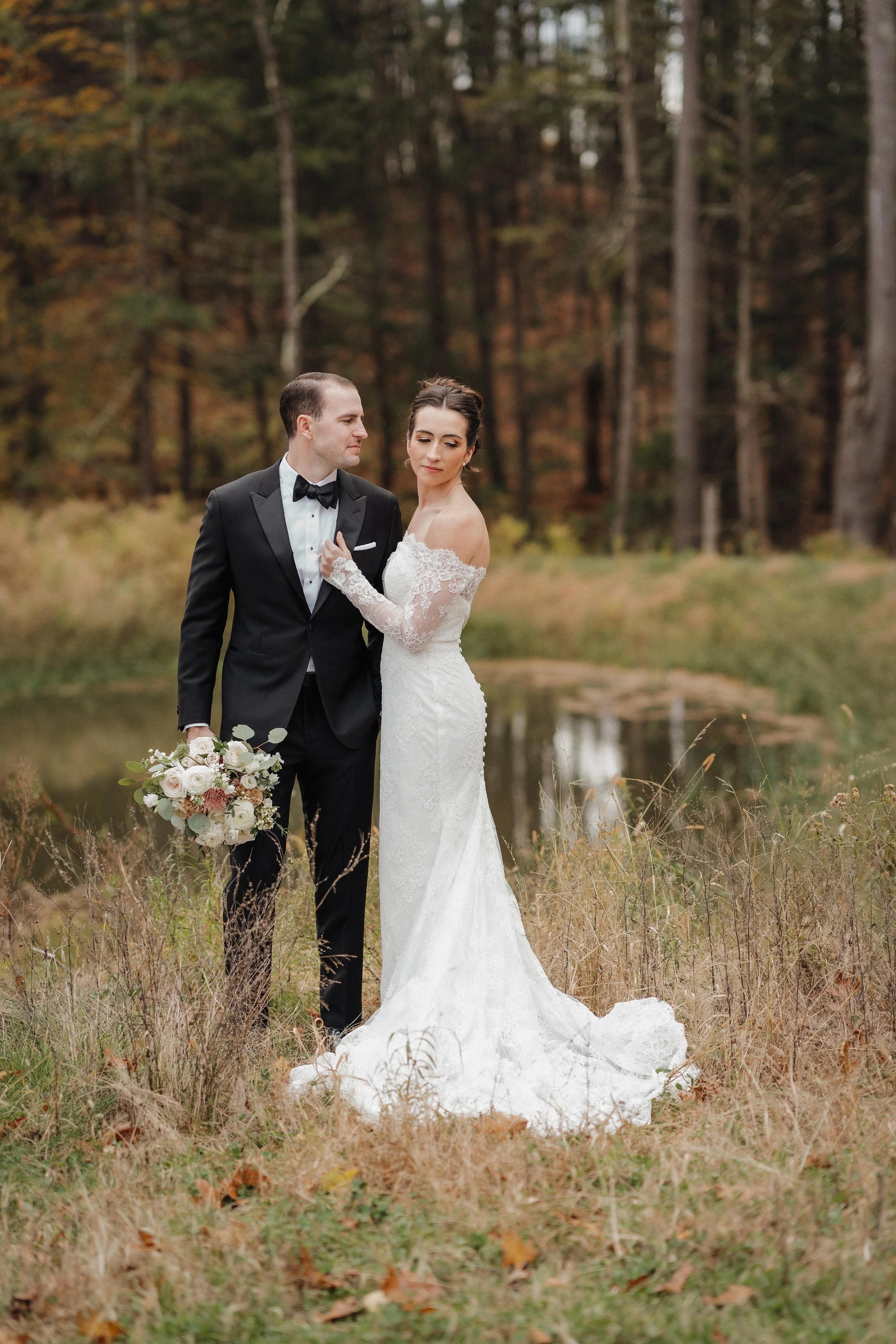 Bride and groom in wedding attire standing outdoors near a pond in a forest with fall foliage.
