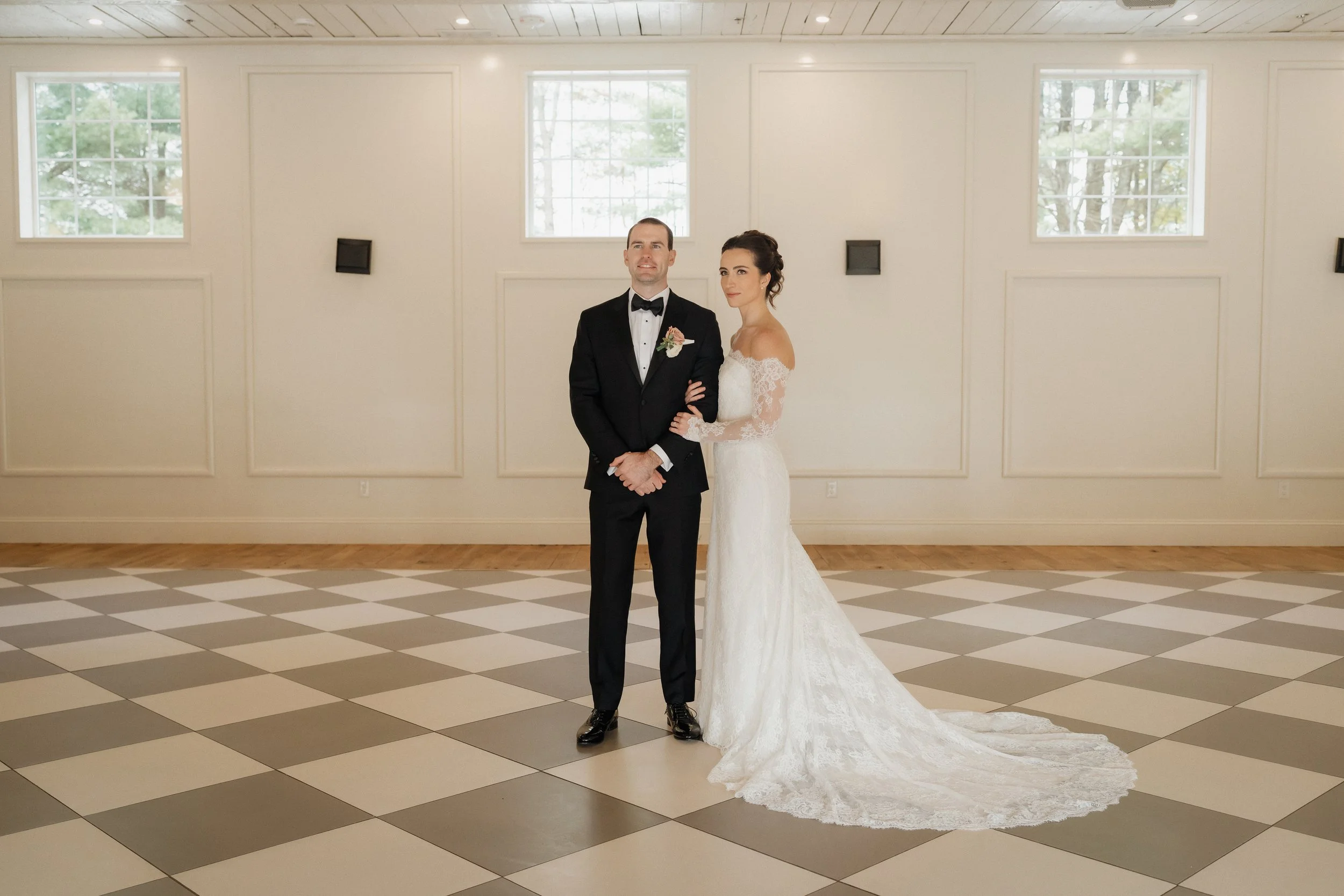 A bride and groom standing arm in arm in a well-lit indoor space with checkered flooring and large windows.