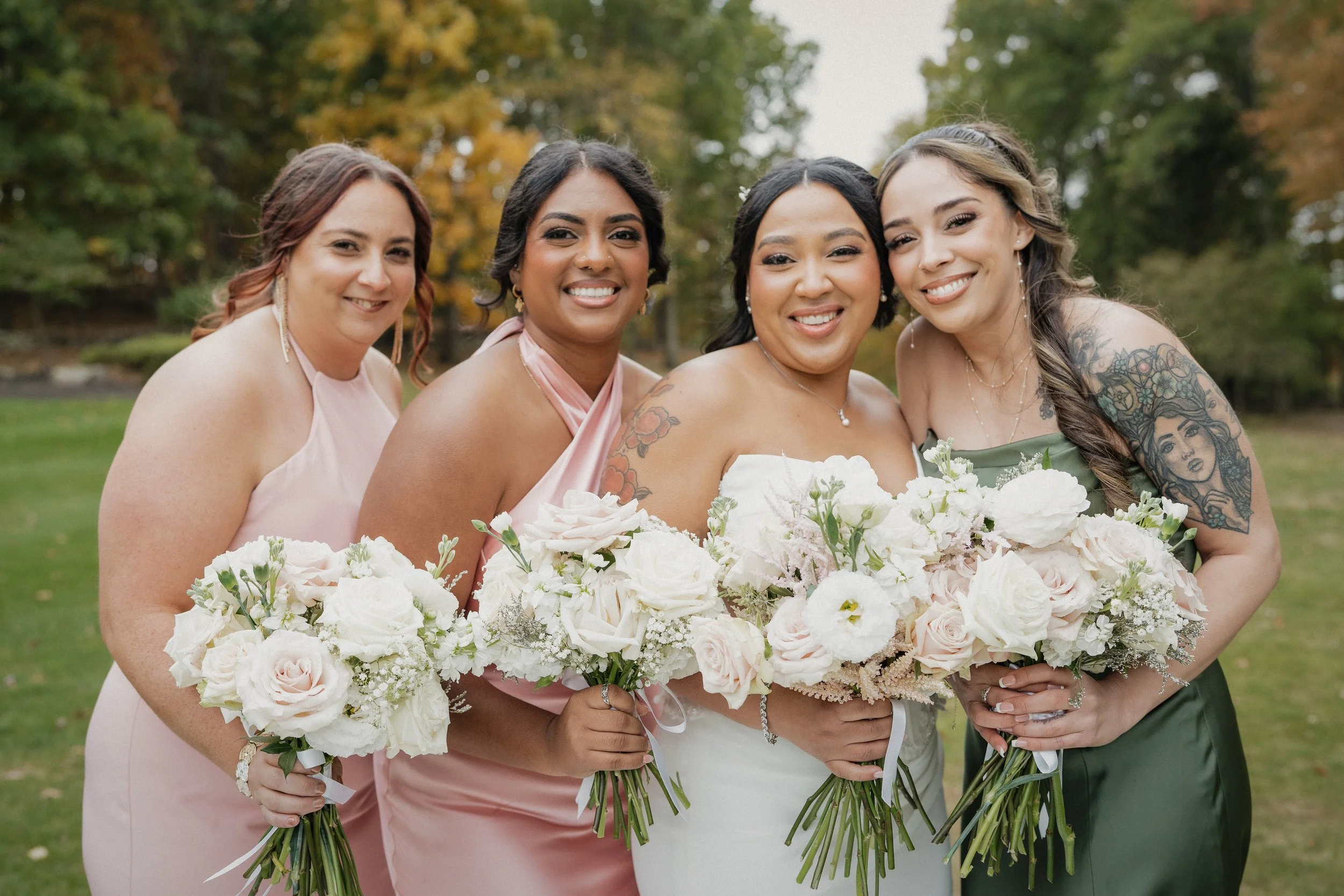 Group of four women dressed in elegant gowns holding bouquets of white and blush roses, standing outdoors on a grassy area with trees in the background.