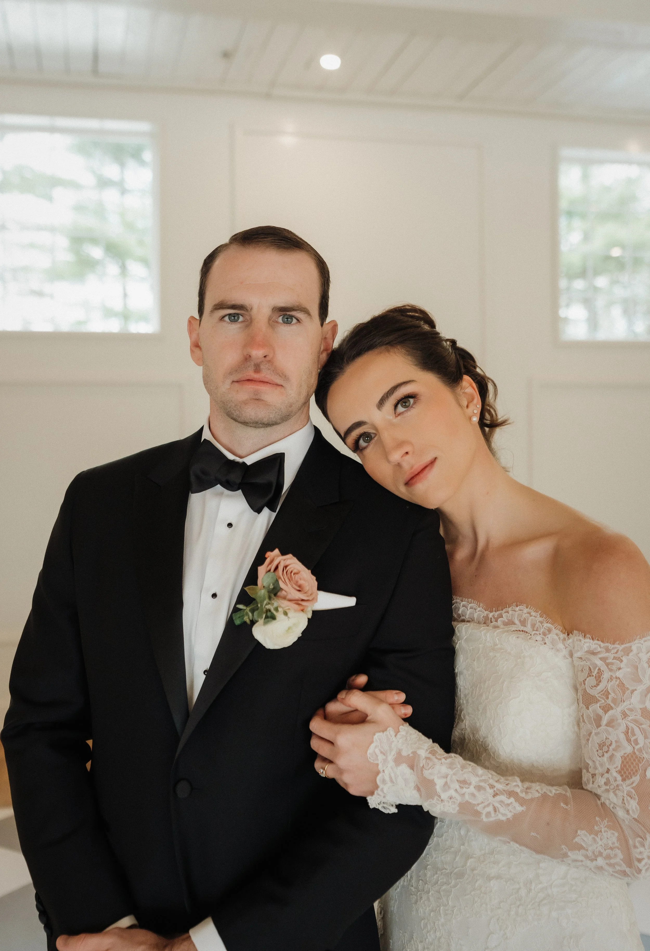 A bride and groom posing indoors, with the bride resting her head on the groom's shoulder, both looking at the camera. The groom is wearing a black tuxedo with a bow tie and a flower boutonniere, and the bride is in a white lace wedding dress with lo