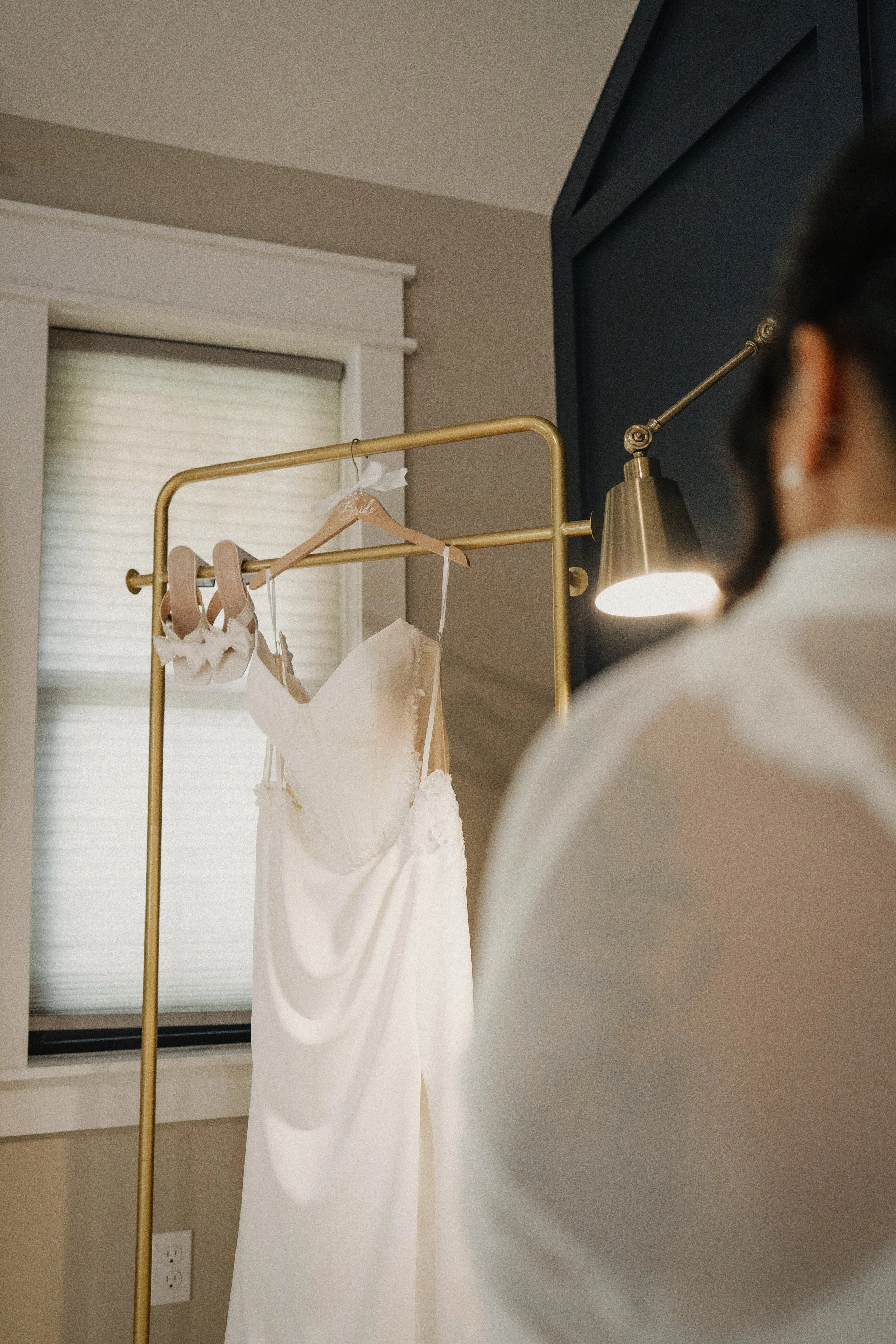 A woman's back as she gazes at a wedding dress hanging on a gold clothing rack in a softly lit room with a window and beige walls.