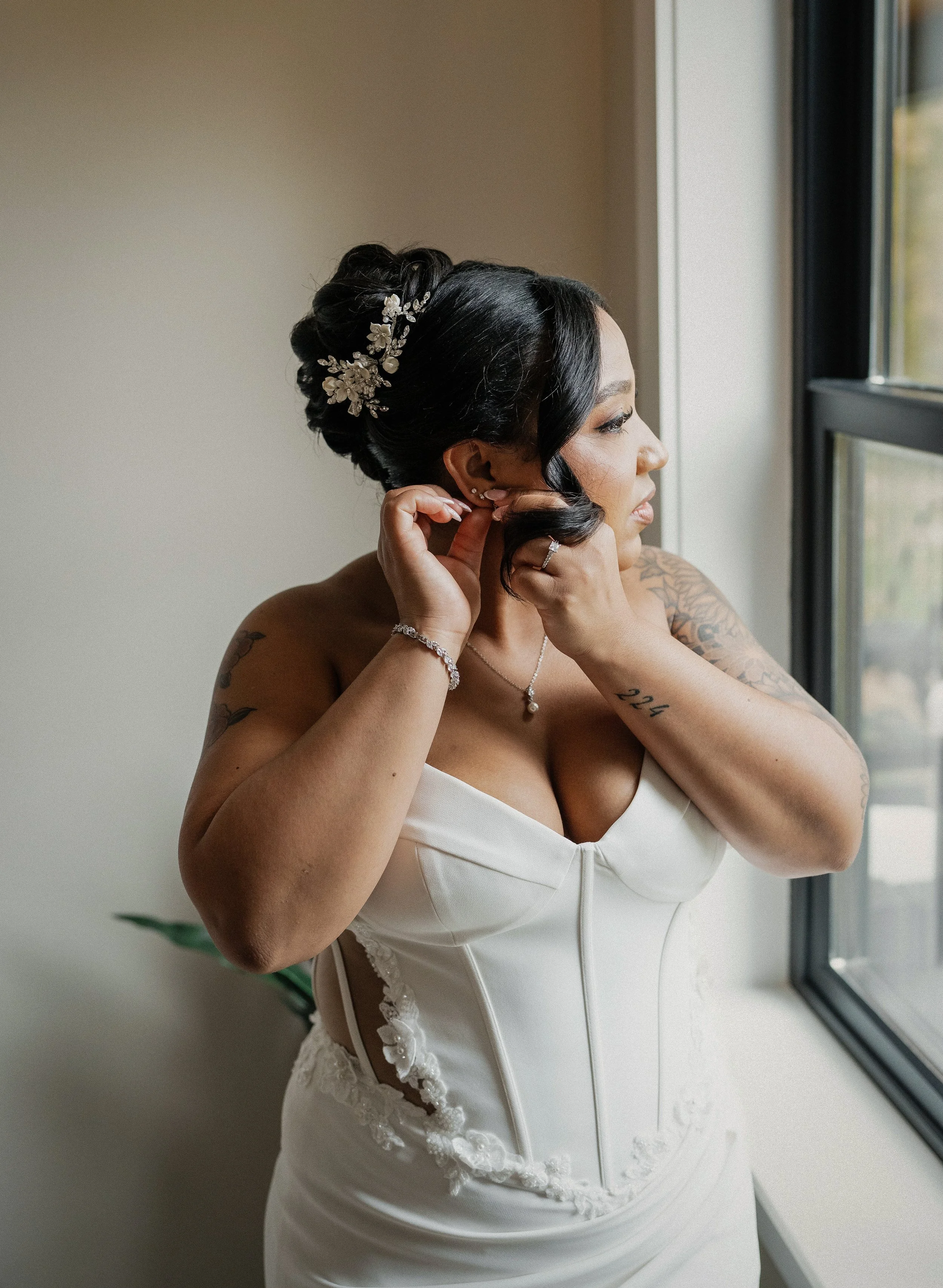 Bride dressed in a white wedding gown, putting on earrings by a window