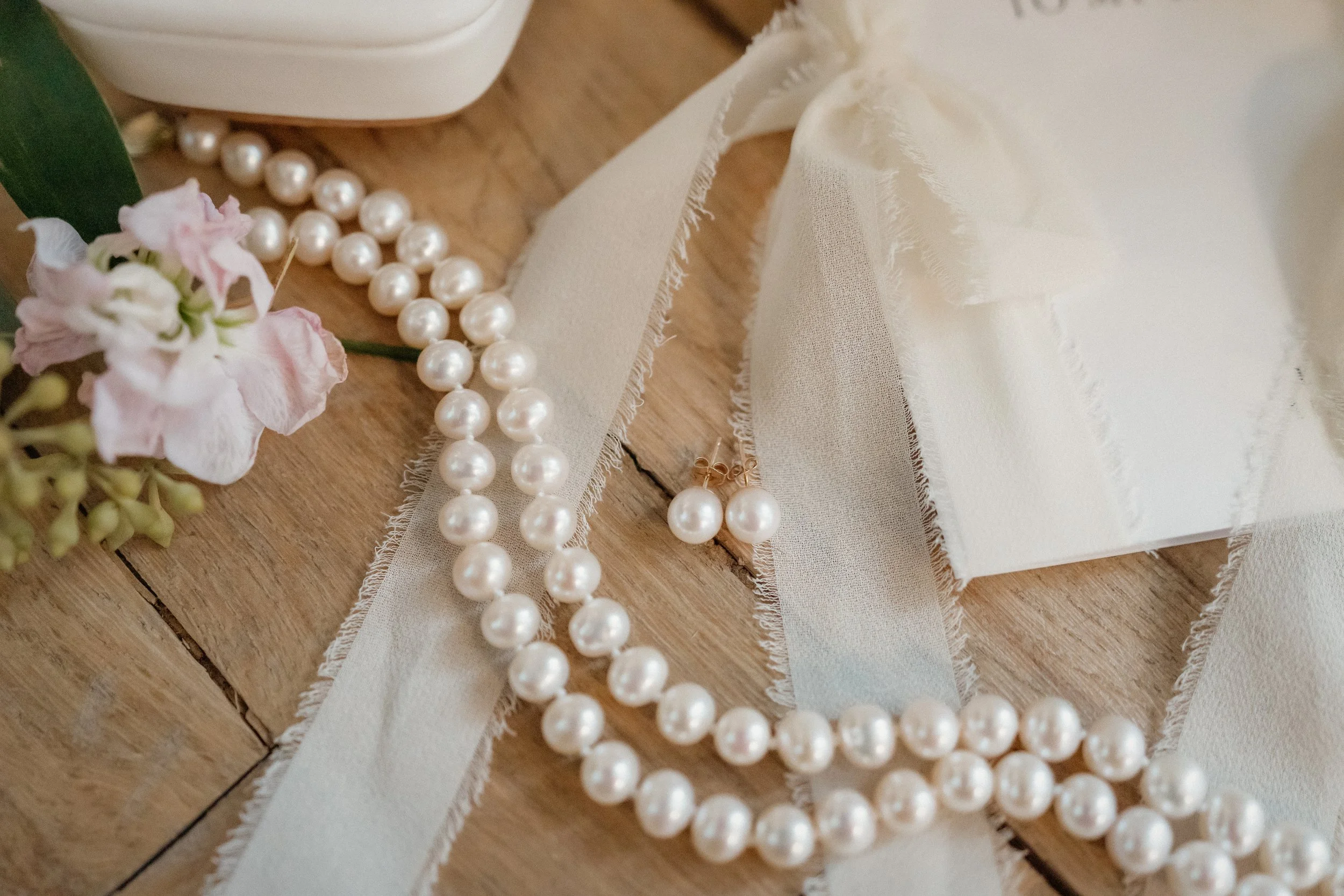 Pearl necklace, pearl earrings, pink flower, ribbon, and gift box on wooden surface.
