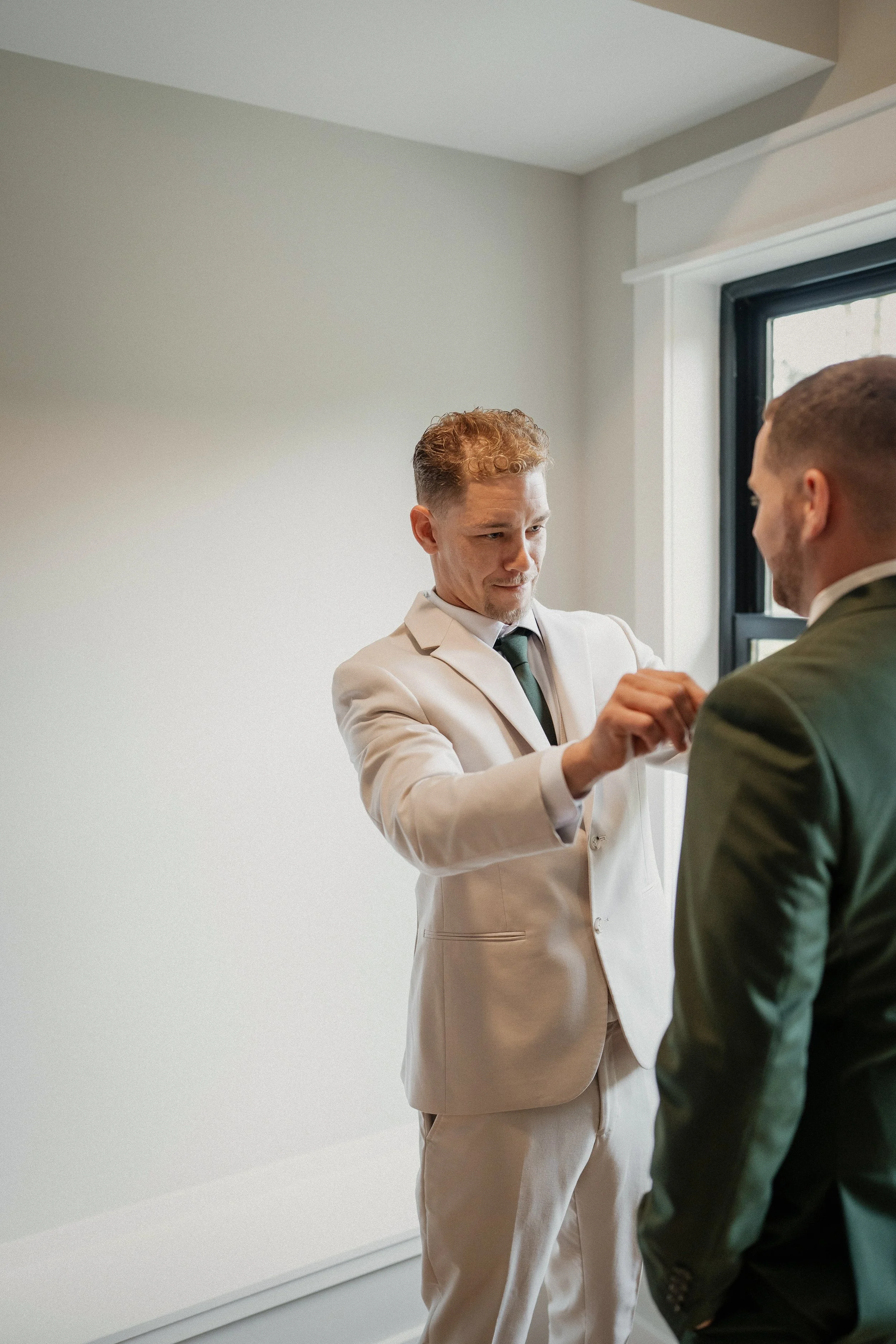 Two men dressed in suits, one pinning a boutonniere on the other's lapel near a window in a minimal decorated room.