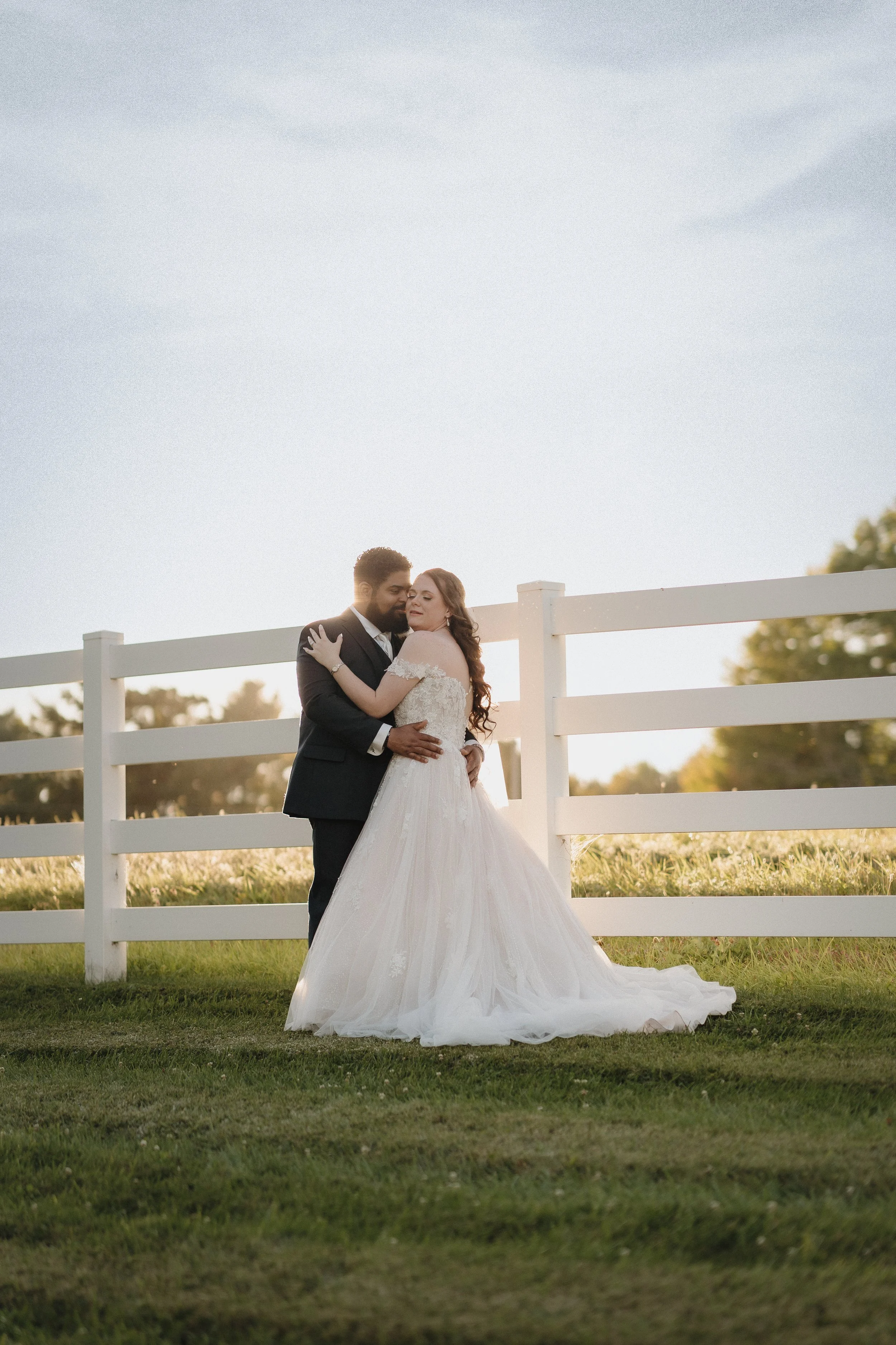 A bride and groom embrace outdoors at sunset, standing on grass next to a white fence, with trees in the background.