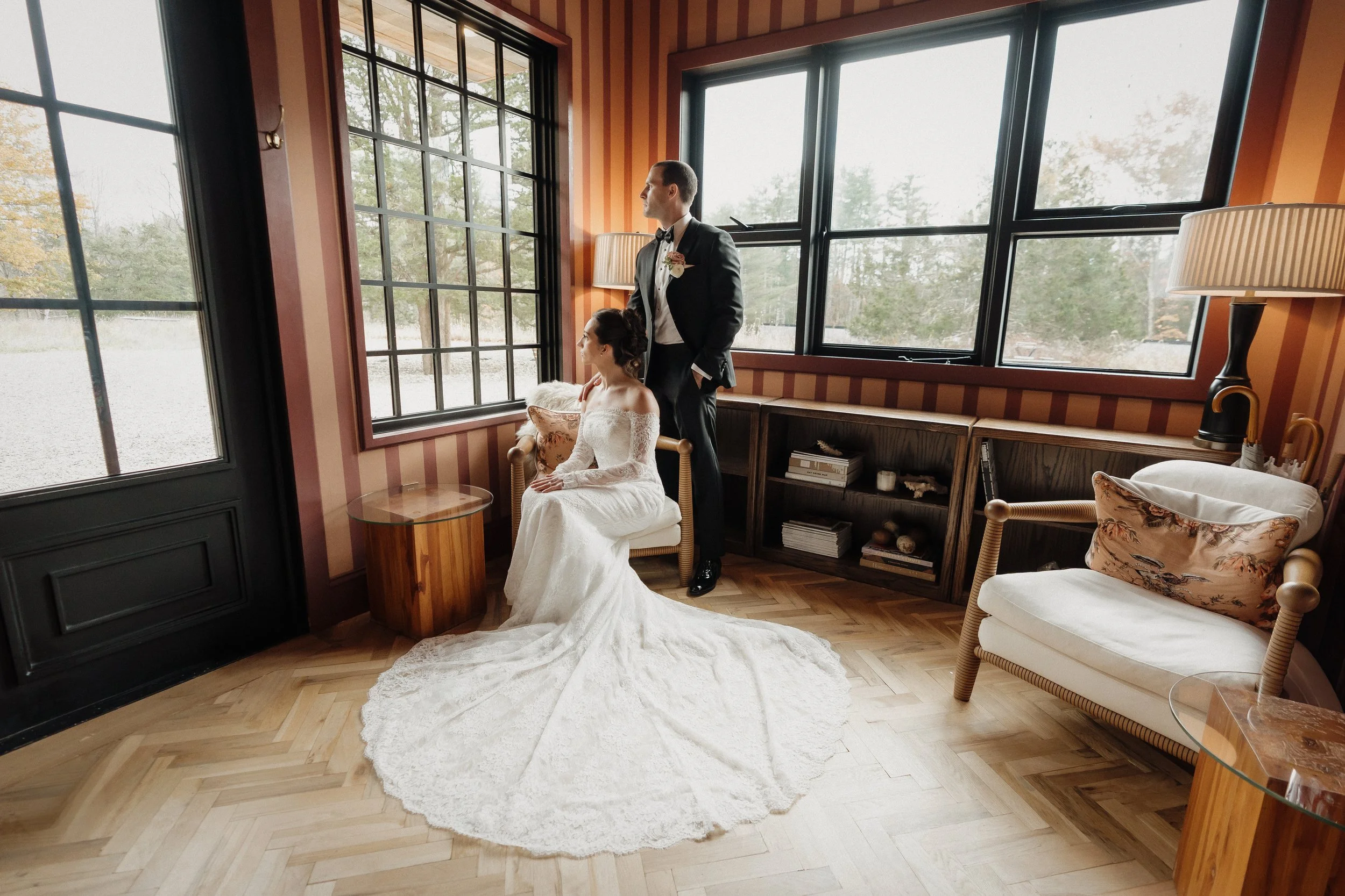 Bride in wedding dress sitting on a chair, groom in tuxedo standing behind her, both inside a room with large windows and striped wallpaper.