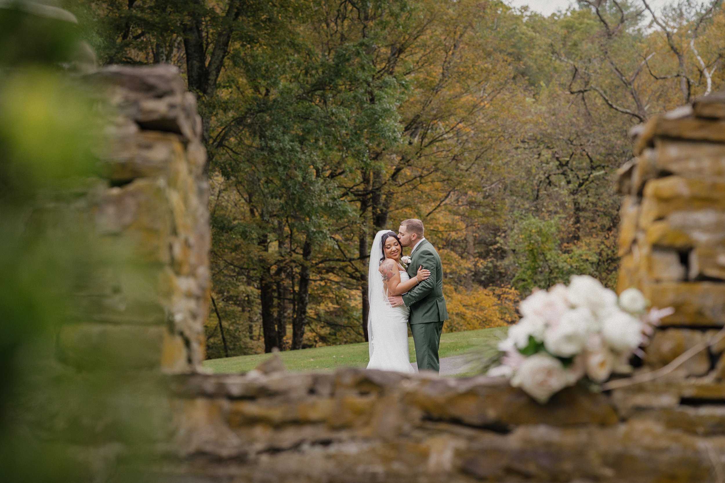 A bride and groom kissing outdoors in a wooded area, seen through a stone opening with a bouquet of white flowers in the foreground.