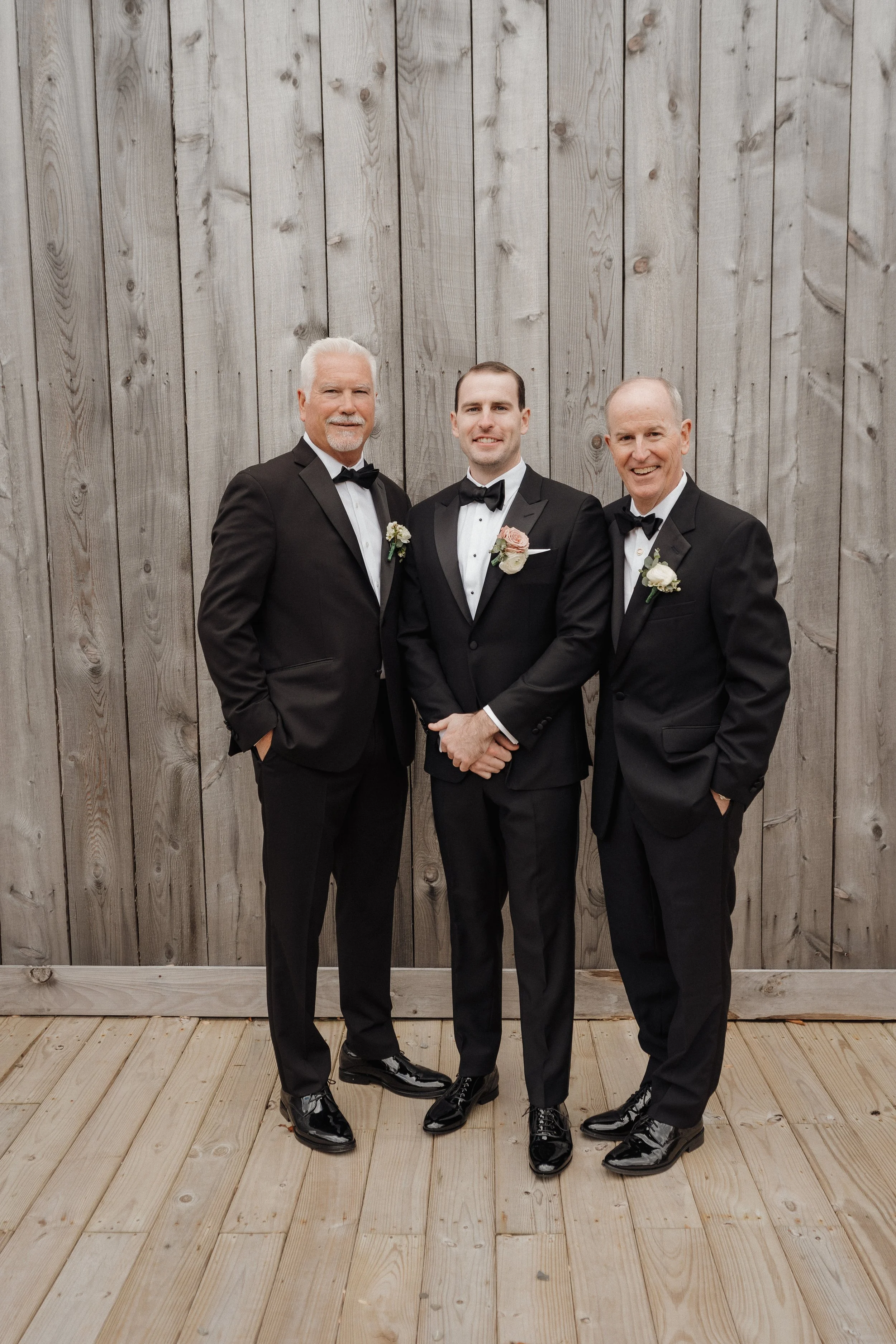 Three men in tuxedos standing side by side in front of a wooden wall, smiling at the camera.