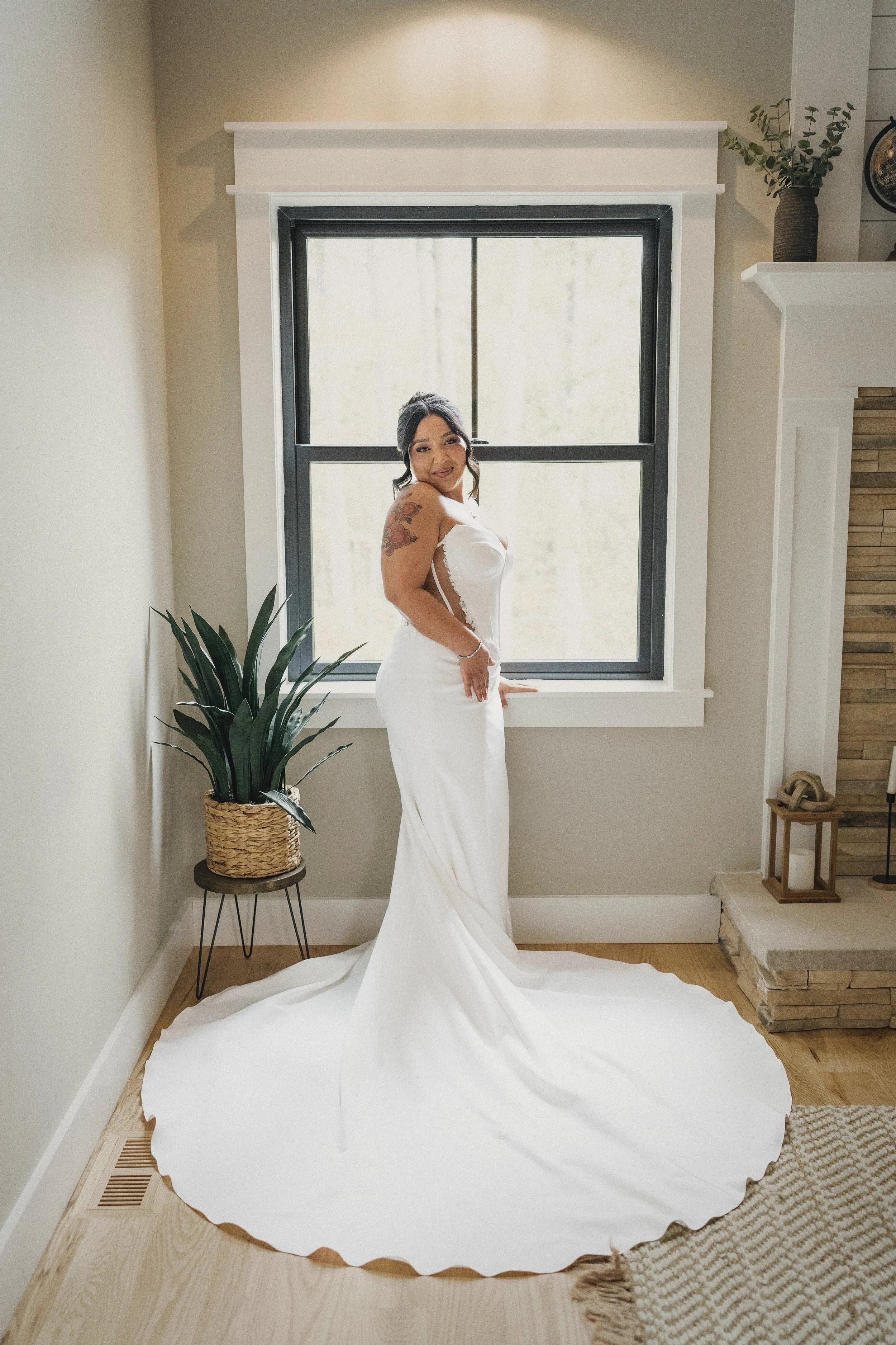 A woman in a white wedding dress standing in front of a window, smiling, with a plant on a stand to her left and a fireplace on her right.