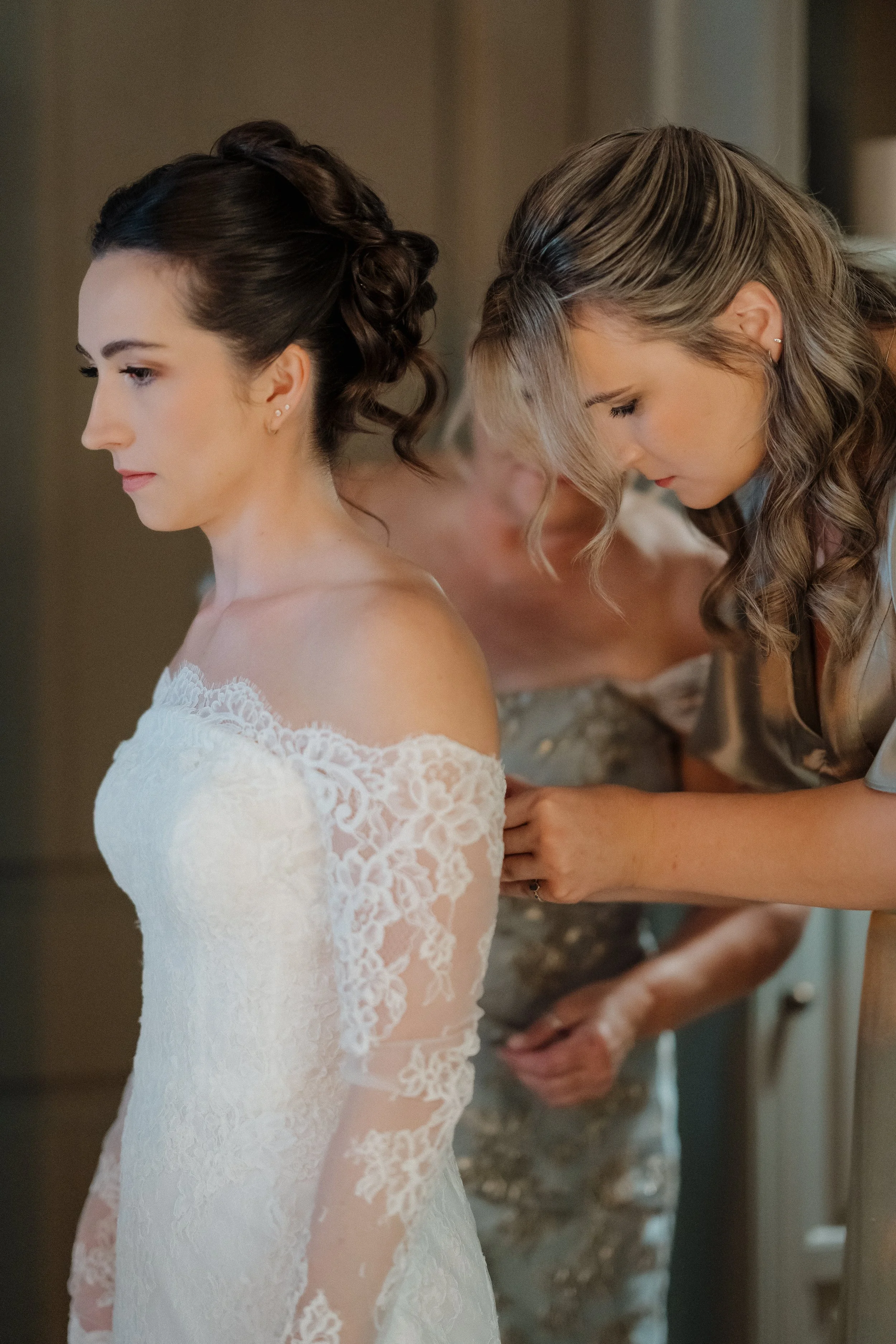 A bride in a lace wedding dress getting help from a woman, likely a bridesmaid or maid of honor, with her gown.