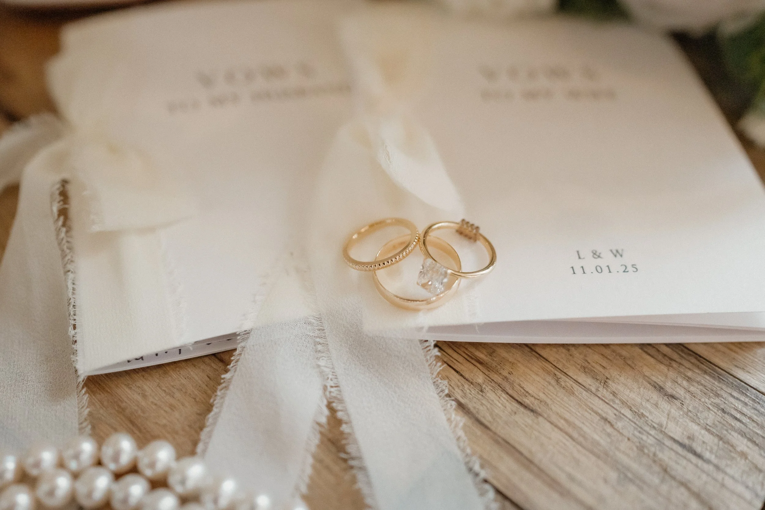 Wedding rings on a white invitation card with a pearl necklace and cream ribbons on a wooden surface.