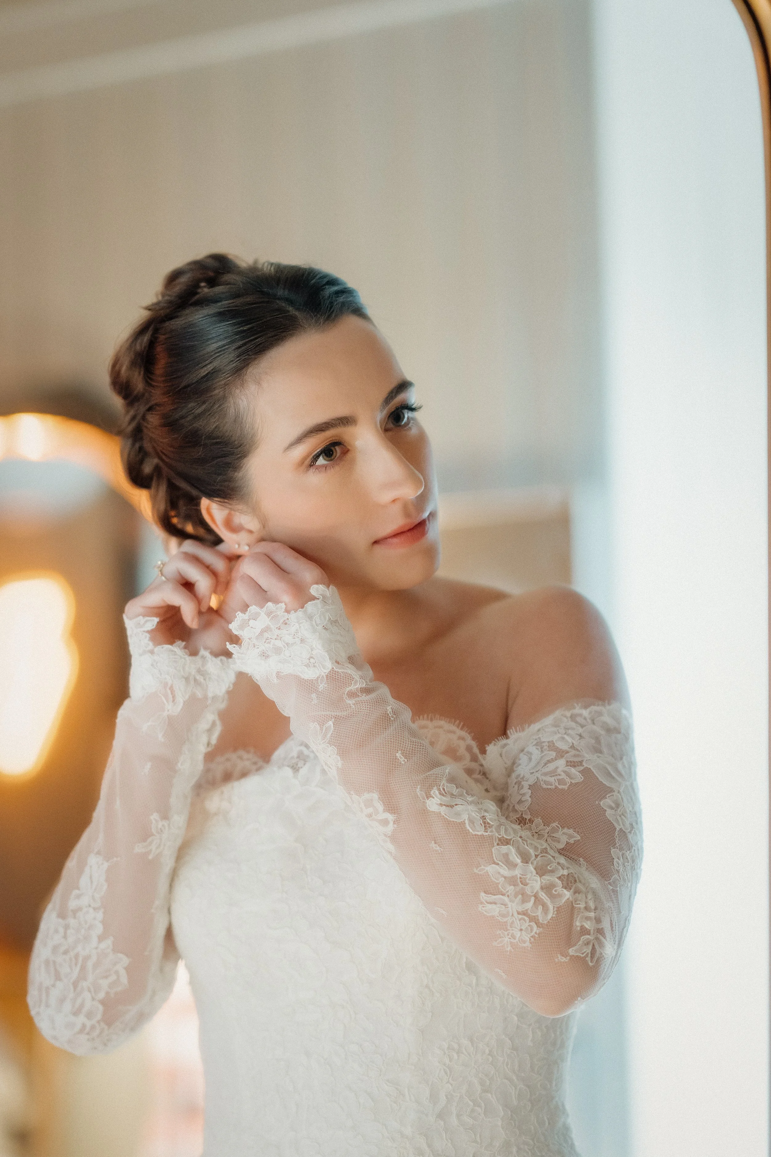 A bride with dark hair in an updo, wearing a lace wedding dress, adjusts her earring in front of a mirror.