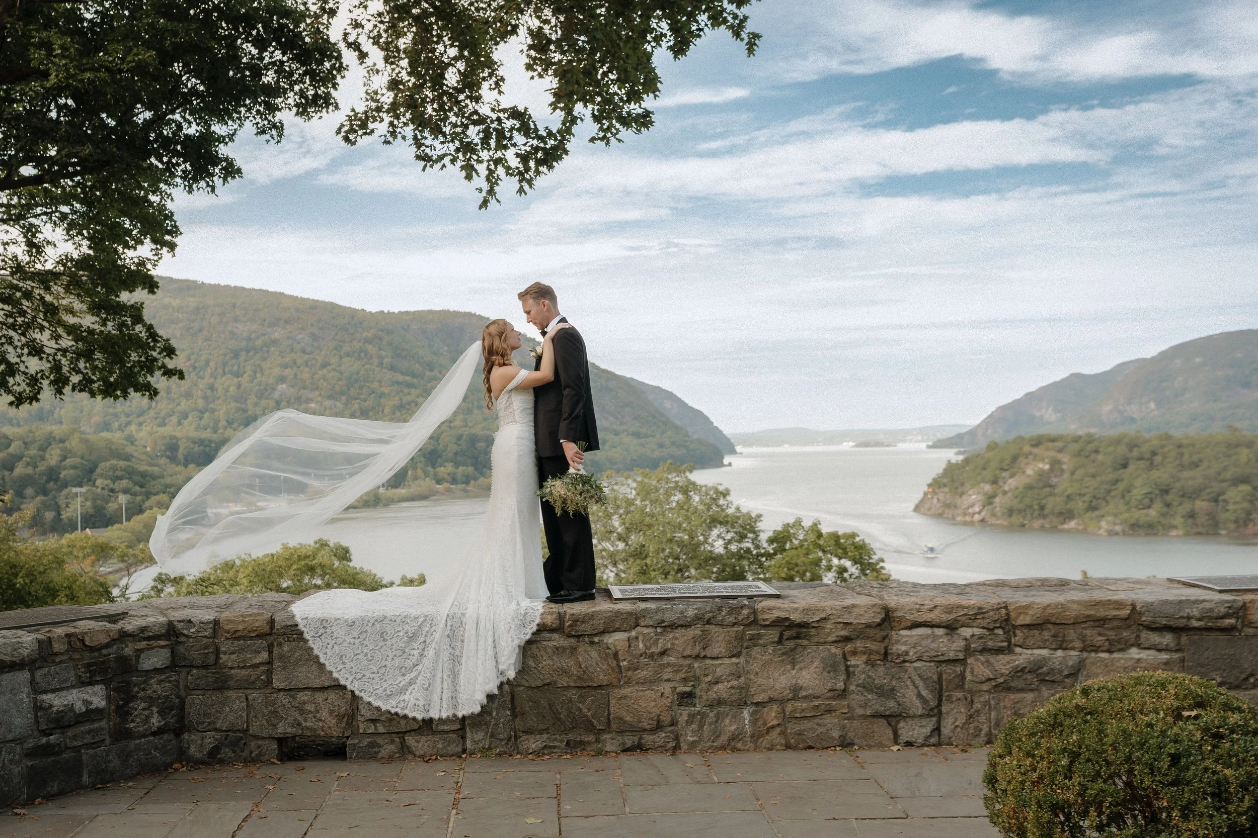 A bride and groom stand close together on a stone ledge overlooking a river with hills in the background. The bride wears a white lace wedding gown with a long train and a flowing veil, while the groom is in a black tuxedo. They gaze into each other's eyes as the bride touches the groom's face, holding a bouquet of flowers in his hand.