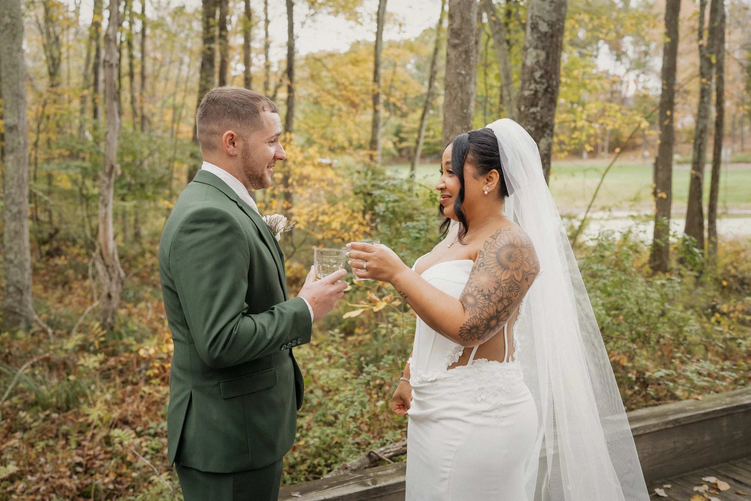 A couple dressed in wedding attire sharing a toast outdoors in a wooded area with fall foliage.