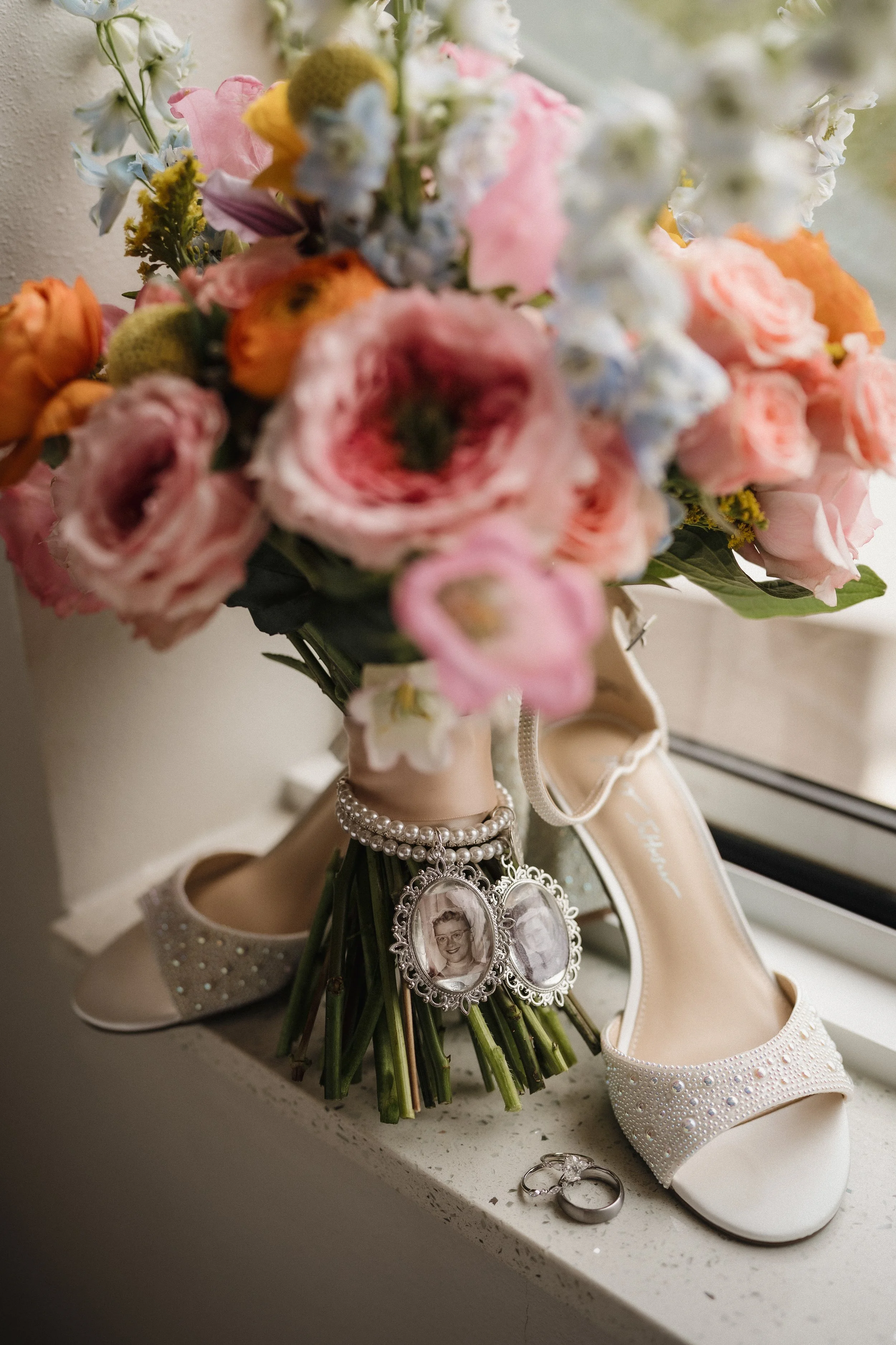 A bouquet of pink, white, and peach flowers placed on a windowsill next to a pair of white bridal shoes with pearl and rhinestone details, a pearl bracelet with photo charms, and a set of wedding rings.