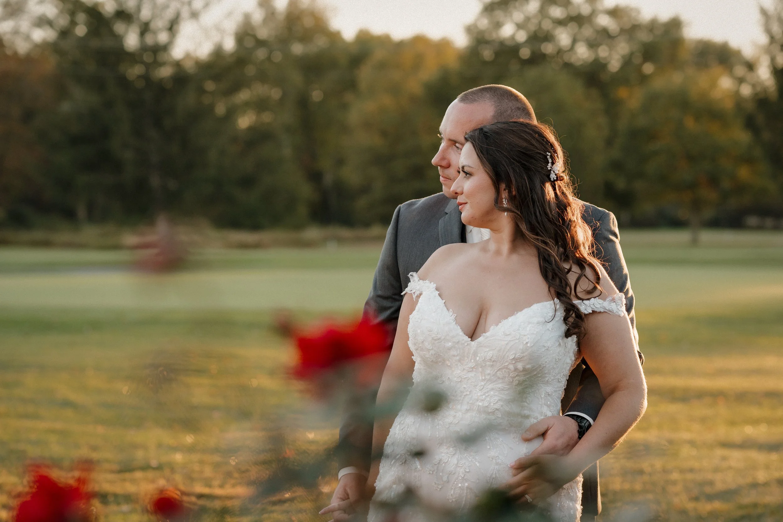 A bride and groom standing together outdoors at sunset, with the bride in a white wedding dress and the groom in a gray suit, surrounded by a grassy field and trees.