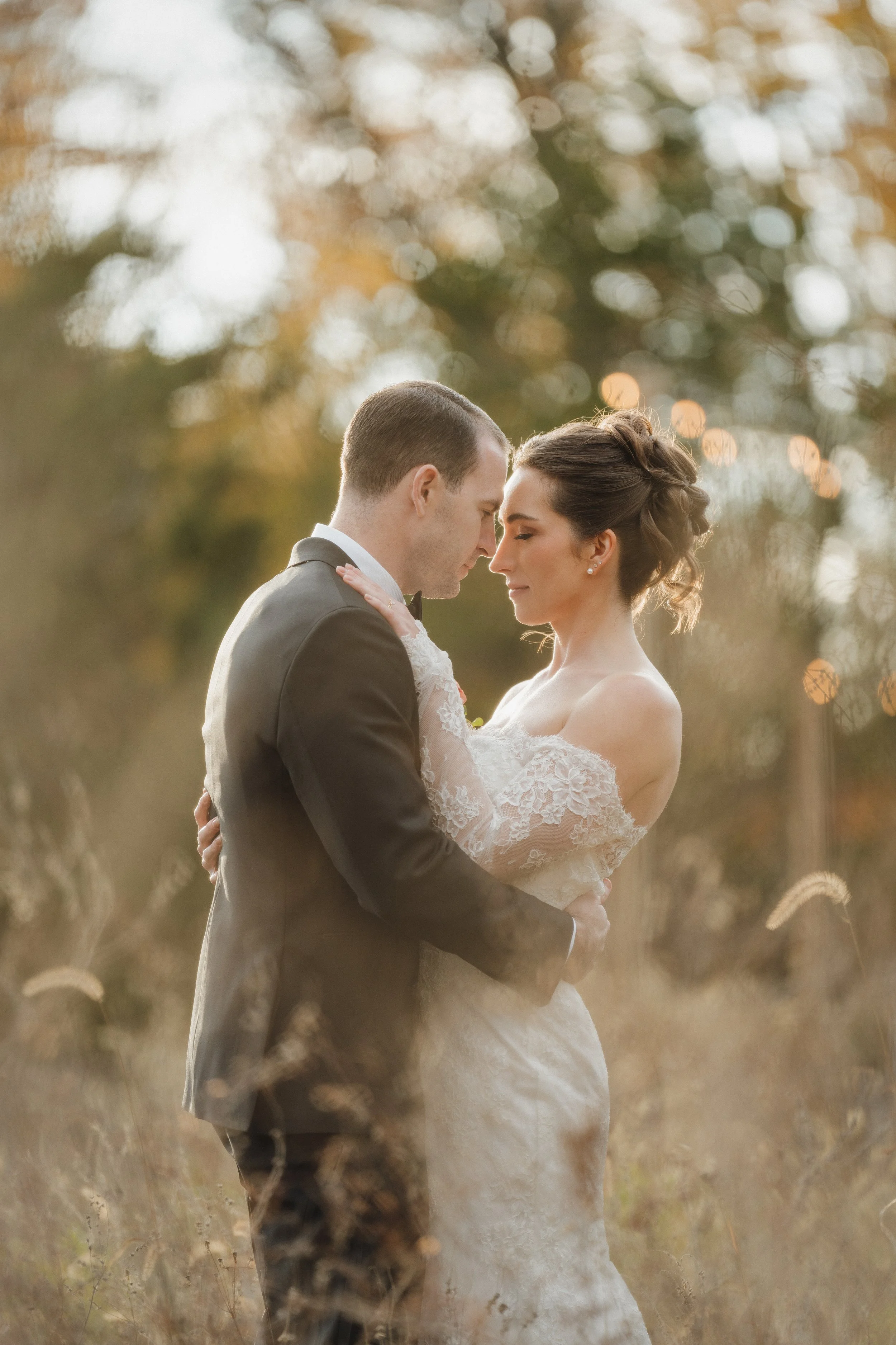 A bride and groom embrace in a field during golden hour, with trees and bokeh lights in the background.