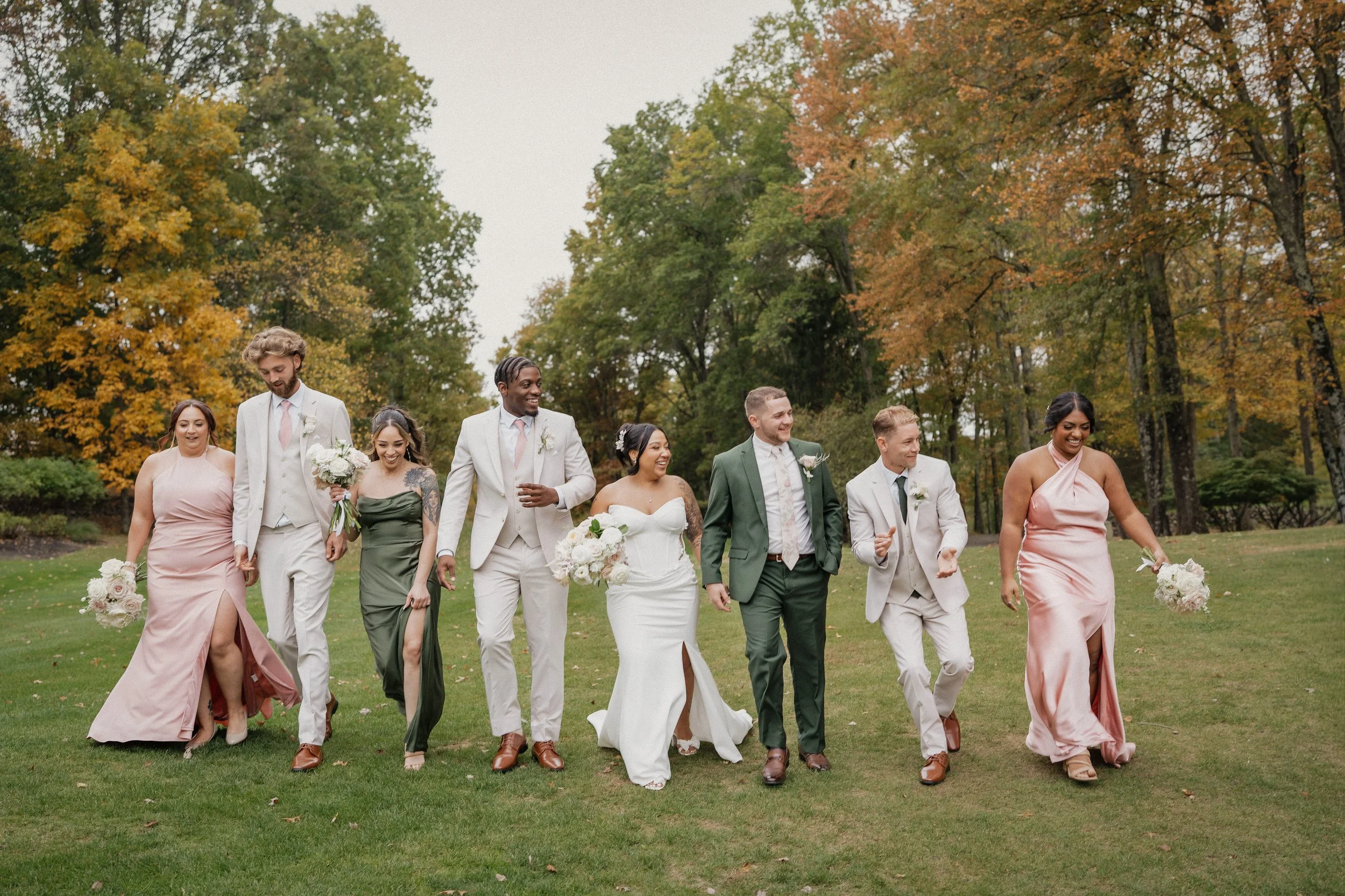 A wedding party walking on grass outdoors with trees displaying fall foliage.